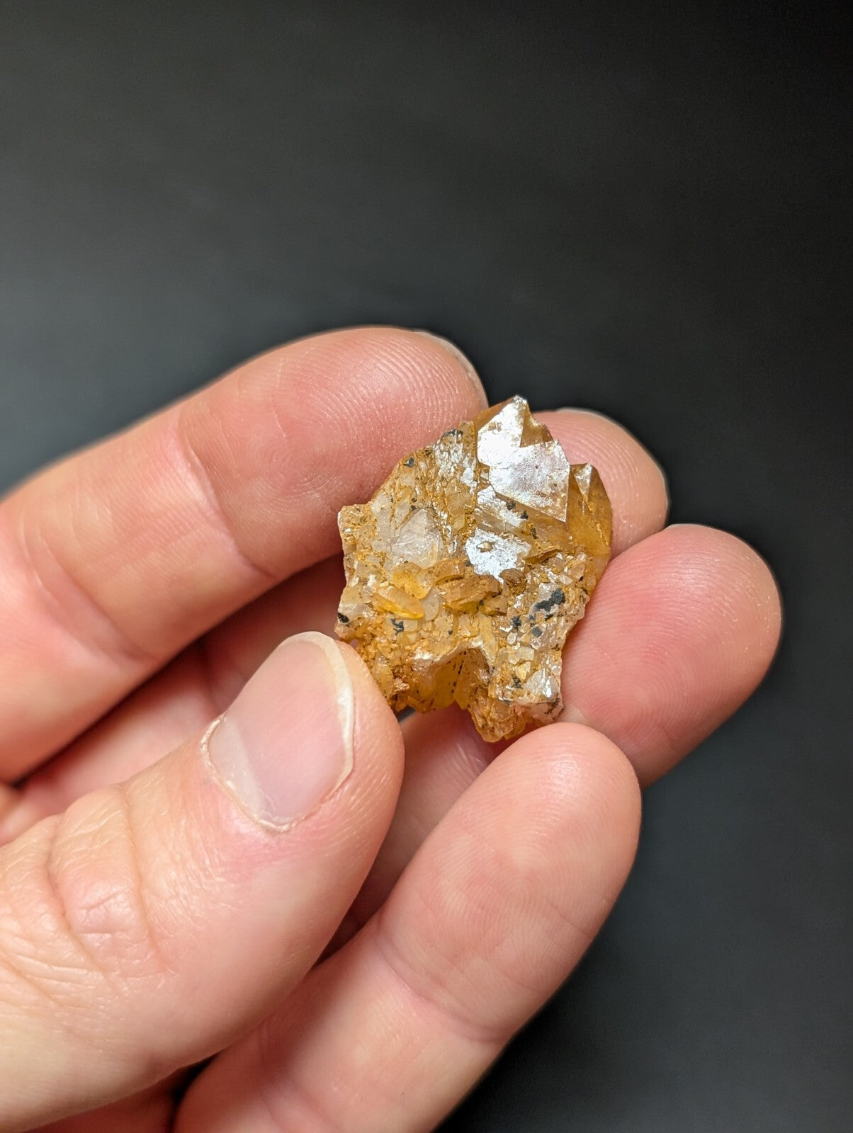 A hand holds a small, irregularly shaped cluster of translucent amber-colored crystals with sharp, faceted edges and some white crystalline inclusions.