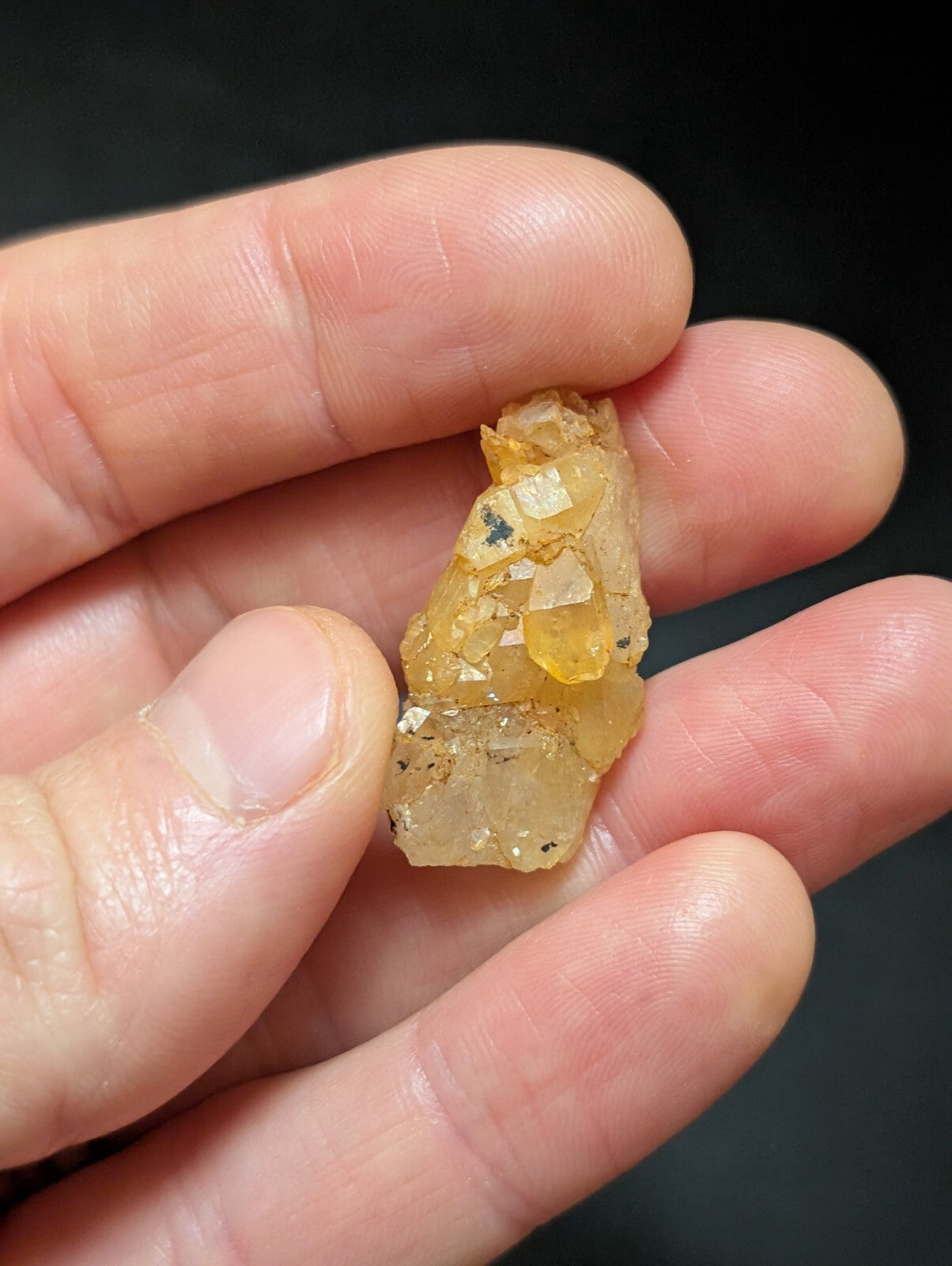 A translucent yellowish-orange mineral specimen with crystalline facets and small dark inclusions, held gently between fingers.