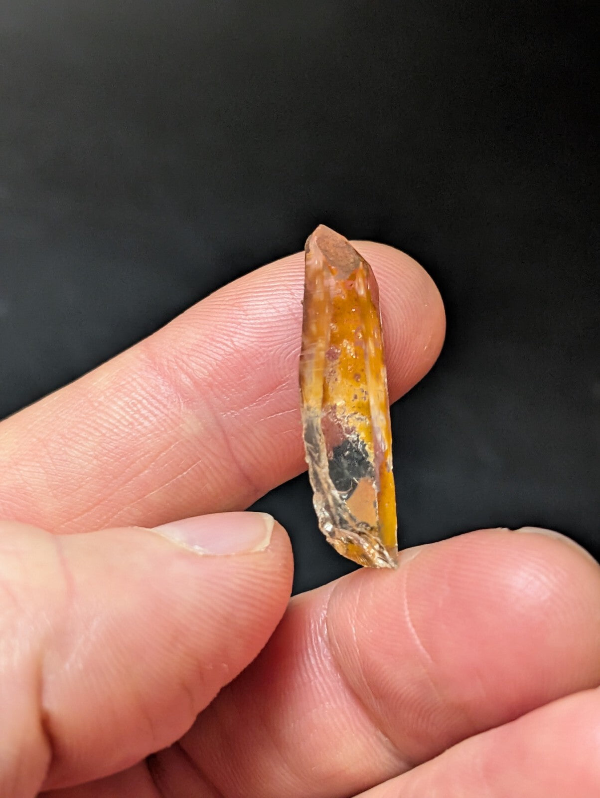 A translucent amber-colored crystal with a pointed tip and natural inclusions, held delicately between fingers.