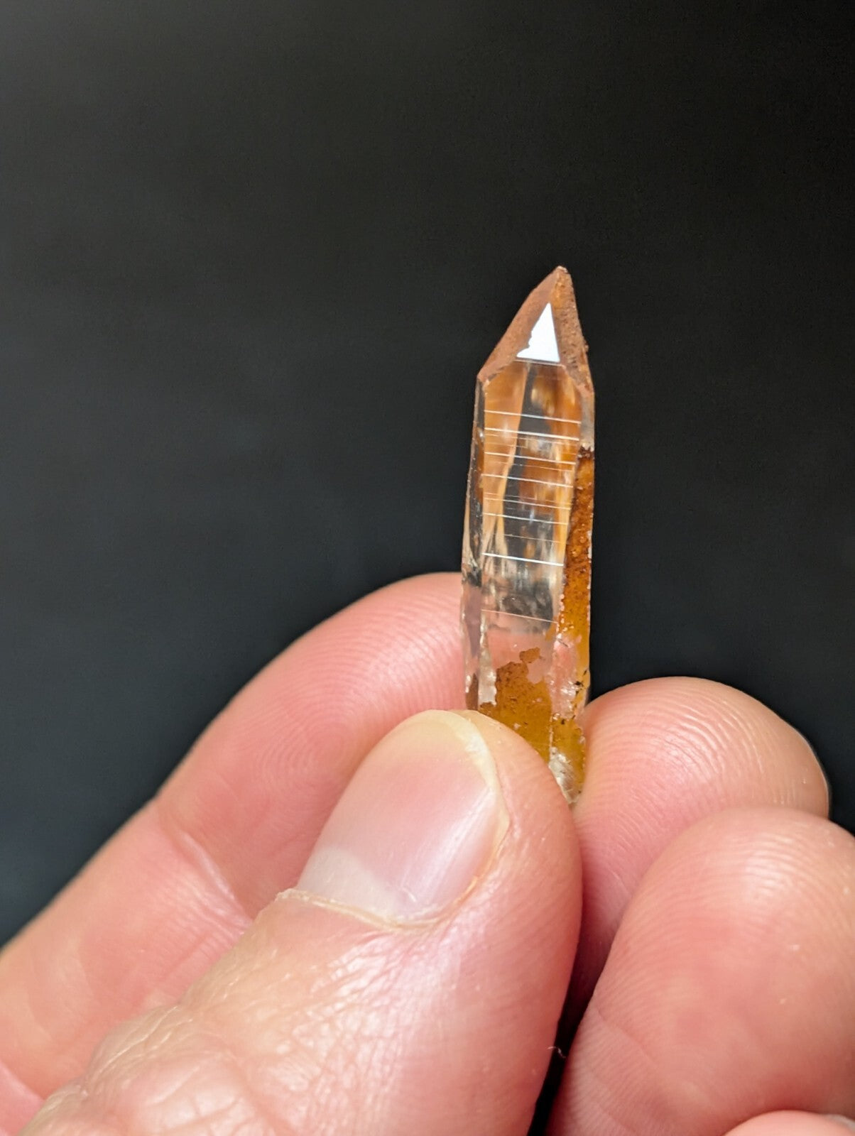 A translucent amber-colored crystal with a pointed tip and visible internal striations, held delicately between fingers.