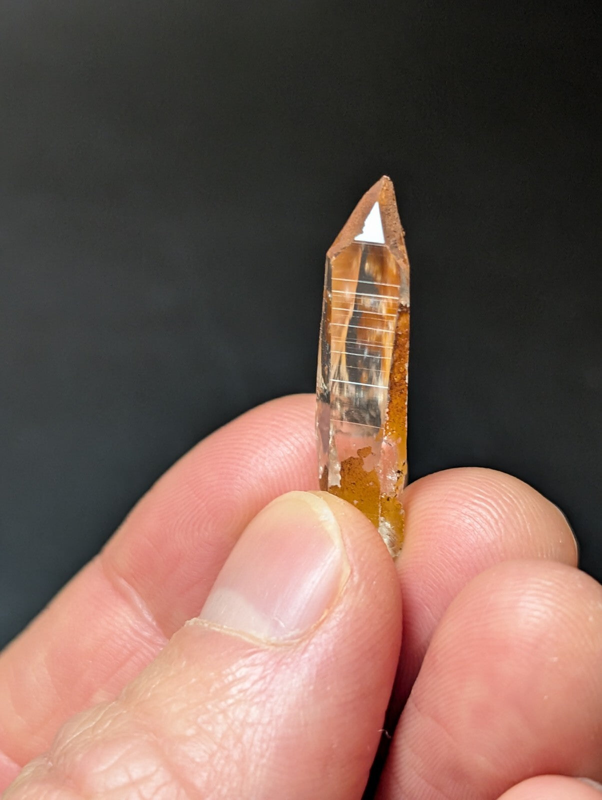 A translucent amber-colored quartz crystal with a pointed tip and visible internal striations, held delicately between fingers.