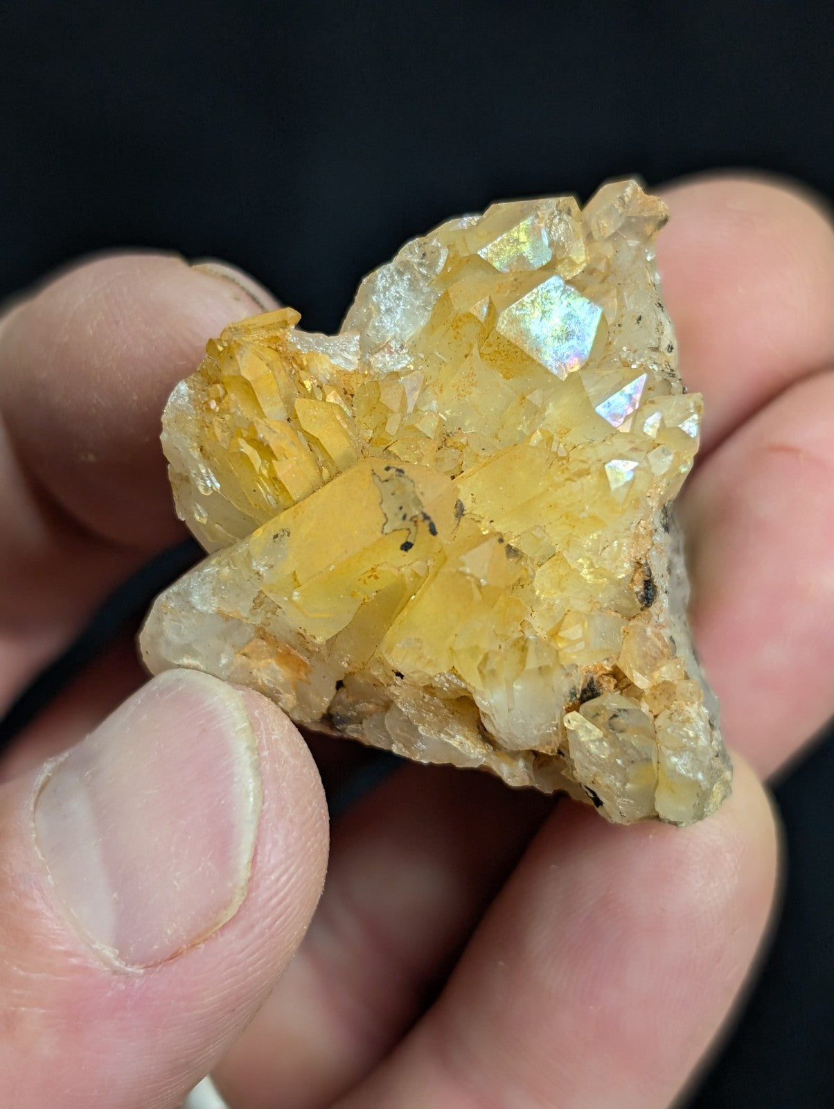 A cluster of translucent yellow crystals with iridescent facets, held in a hand against a dark background.