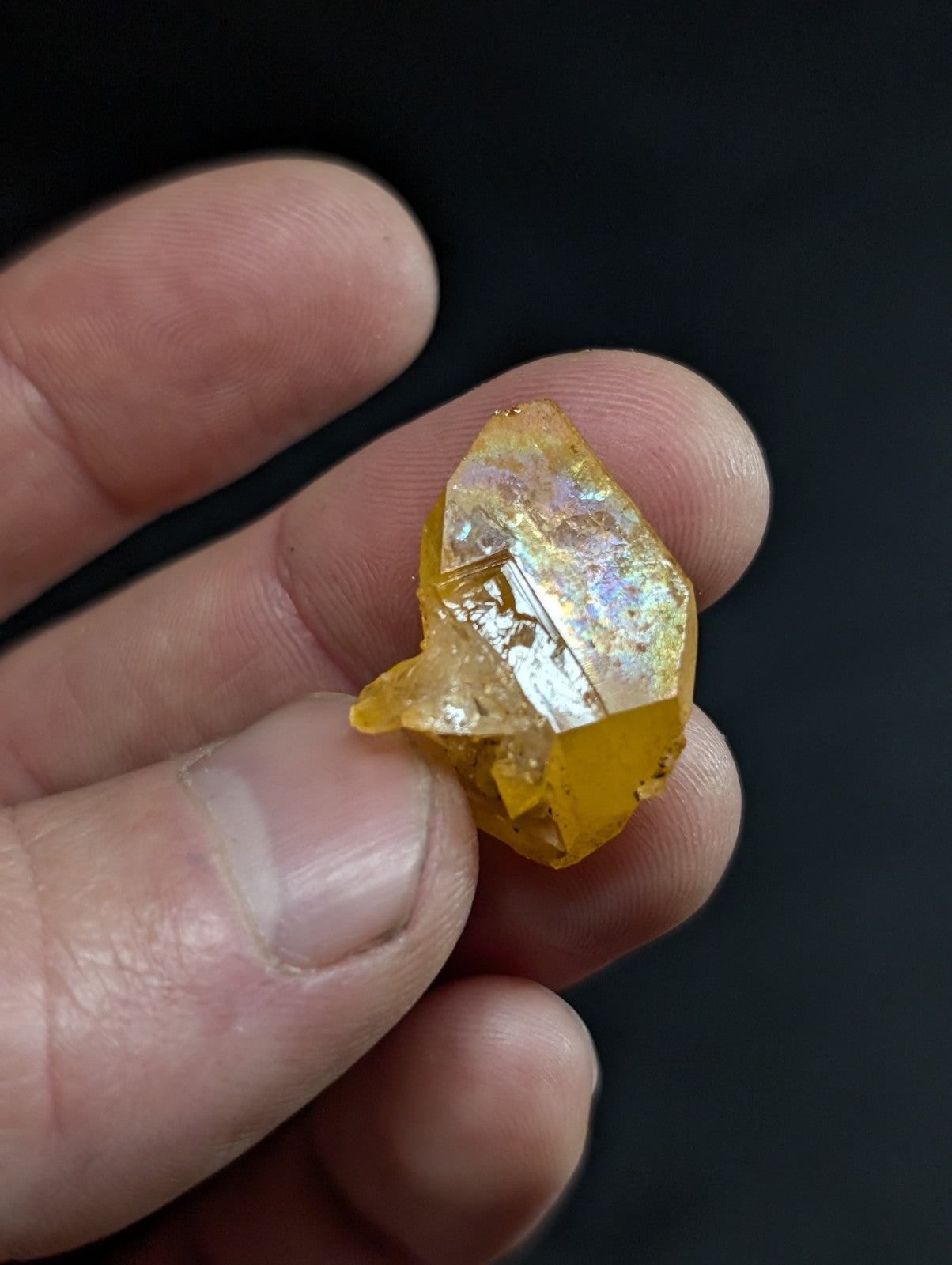 A translucent golden-yellow crystal with iridescent facets, held delicately between fingers.