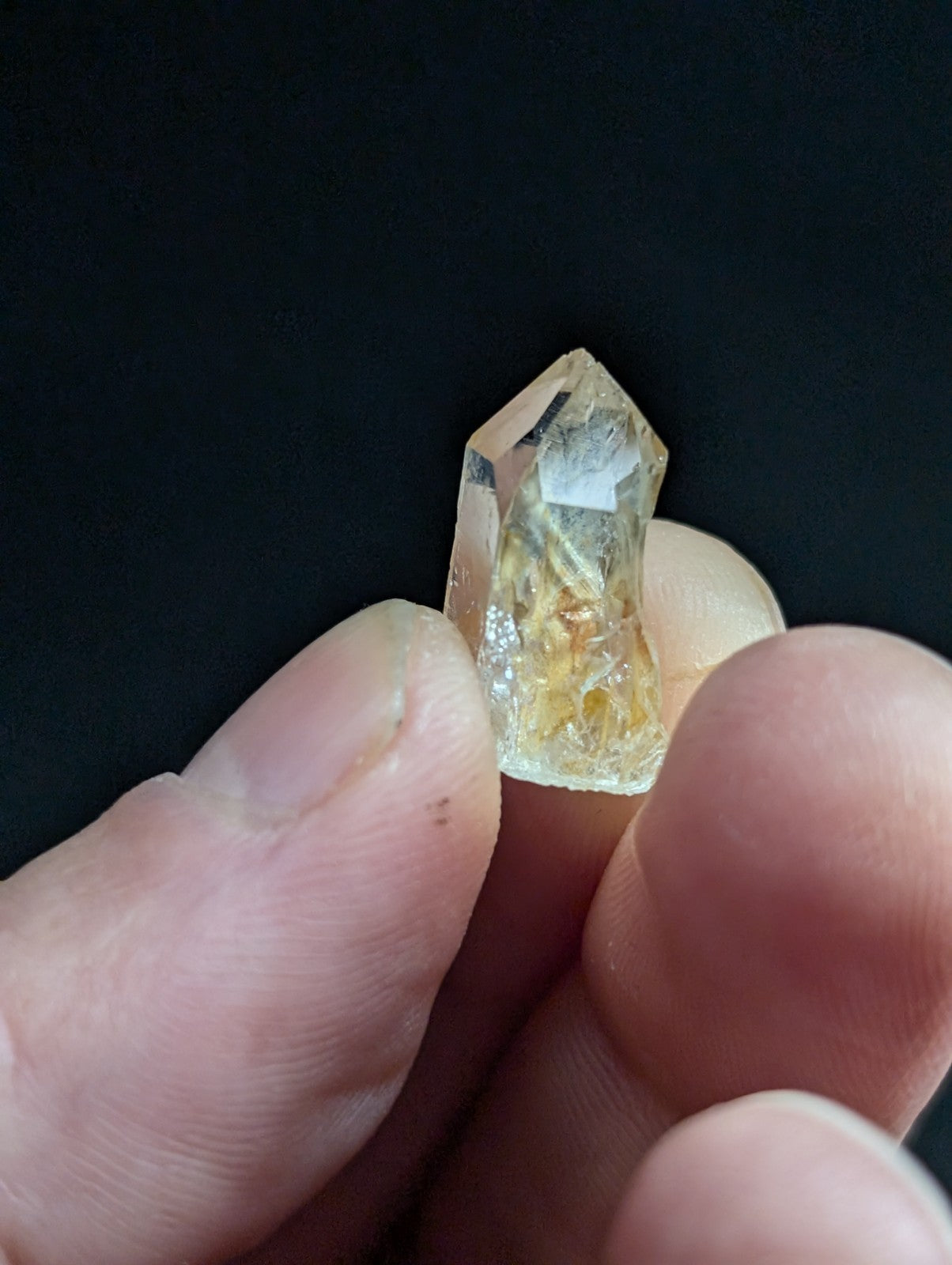 A translucent yellowish crystal with a pointed top and visible internal inclusions, held between fingers.
