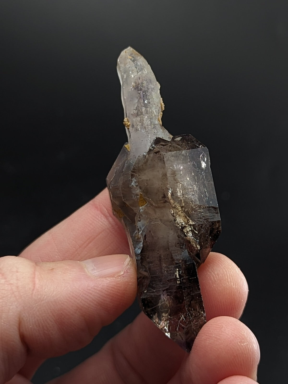 A translucent smoky quartz crystal with a pointed tip and natural inclusions, held gently between fingers against a dark background.