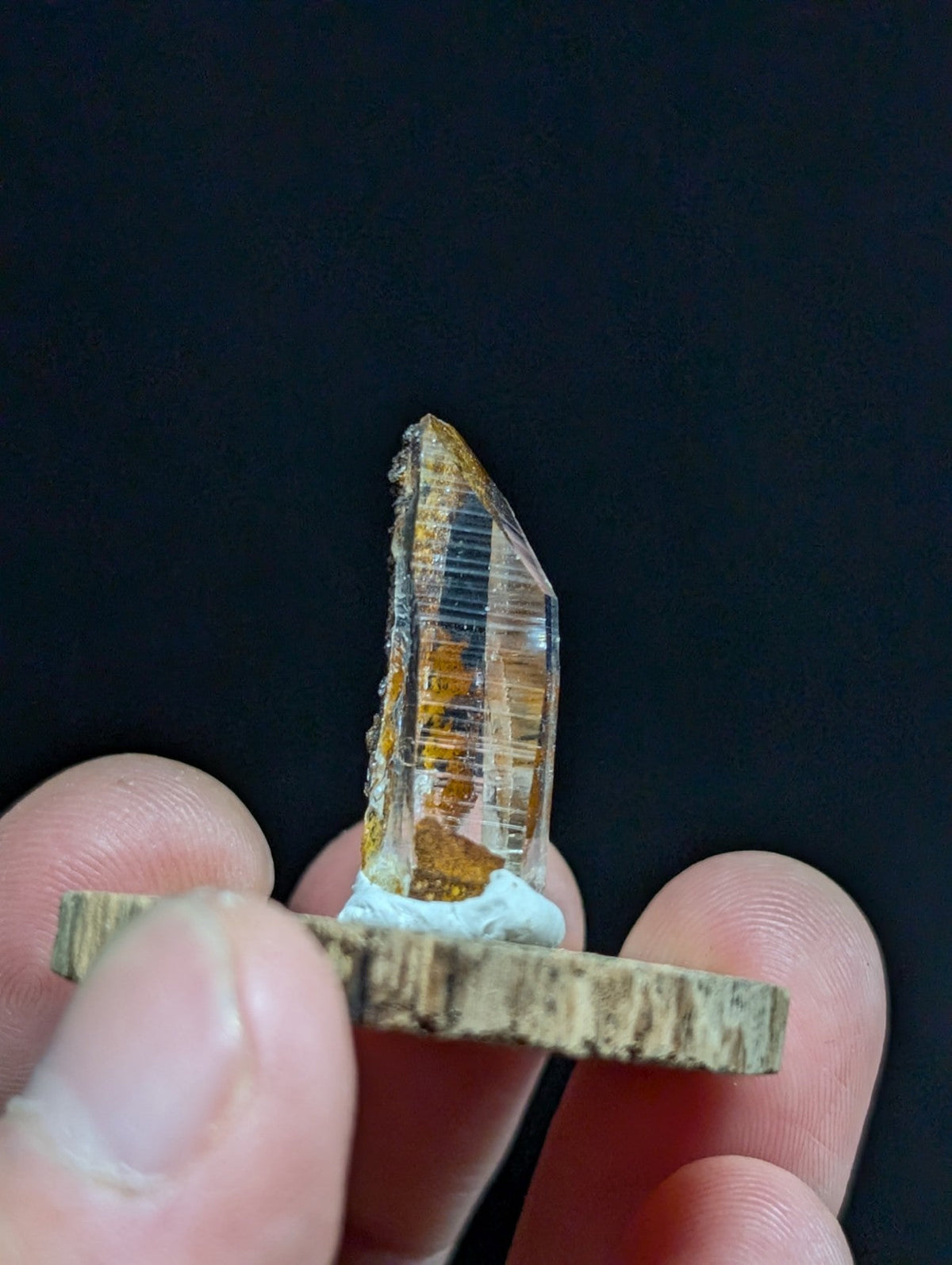 A translucent amber-colored crystal with visible internal striations and a rough, jagged top edge is held upright on a rustic wooden base.
