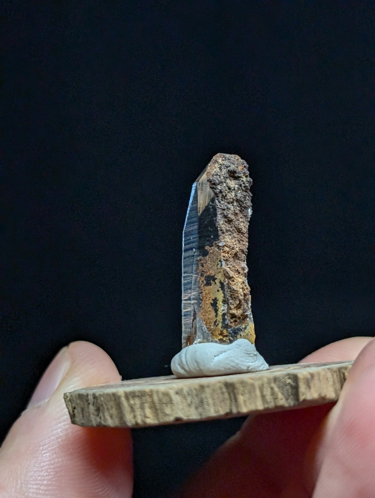 A translucent, vertically oriented quartz crystal with a rough, earthy brown and orange matrix along its upper portion stands upright on a small wooden base.