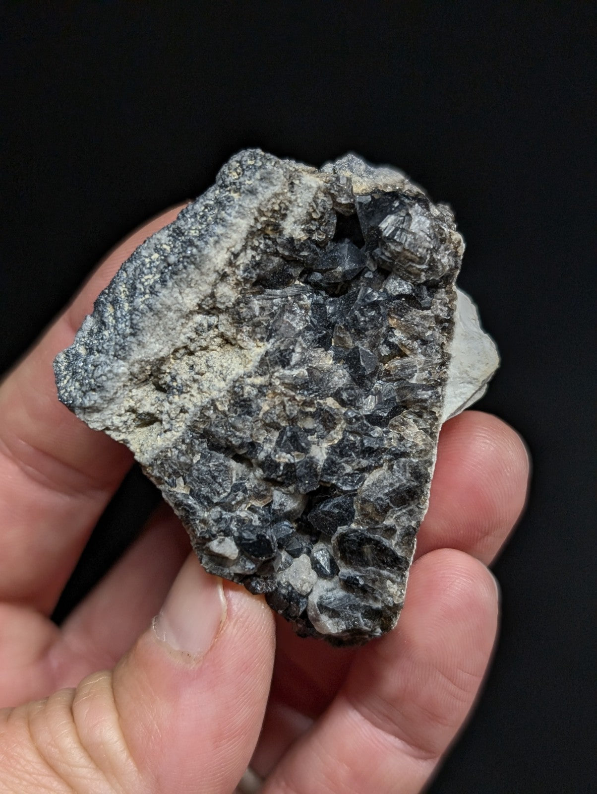 A hand holds a rough, irregularly shaped mineral specimen with dark gray and black crystalline formations against a stark black background.
