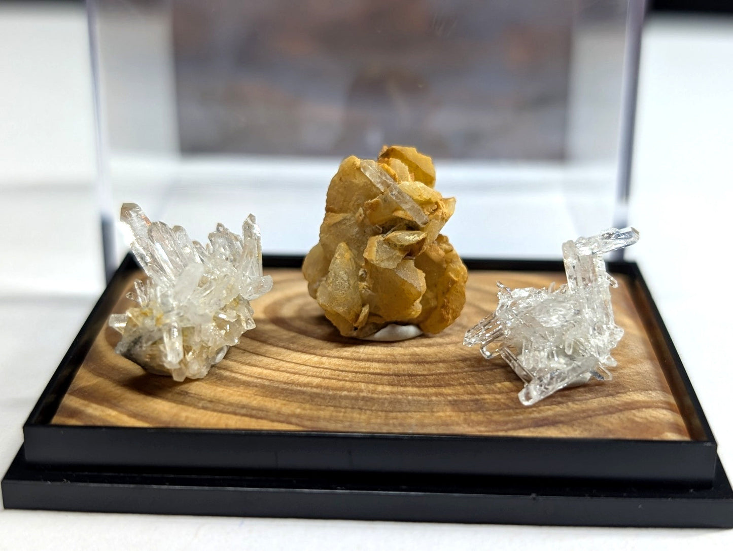 A clear, crystalline quartz specimen with sharp, angular facets sits beside a golden-yellow mineral cluster and another transparent crystal formation on a wooden display tray.