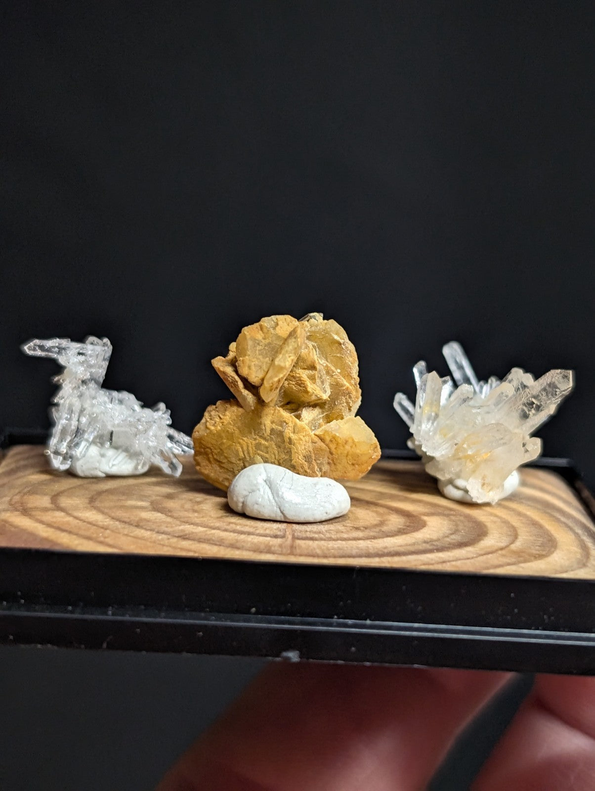 A miniature crystal and mineral display featuring a clear quartz figurine, a golden-yellow rock specimen with layered textures, a smooth white pebble, and another cluster of transparent crystals.