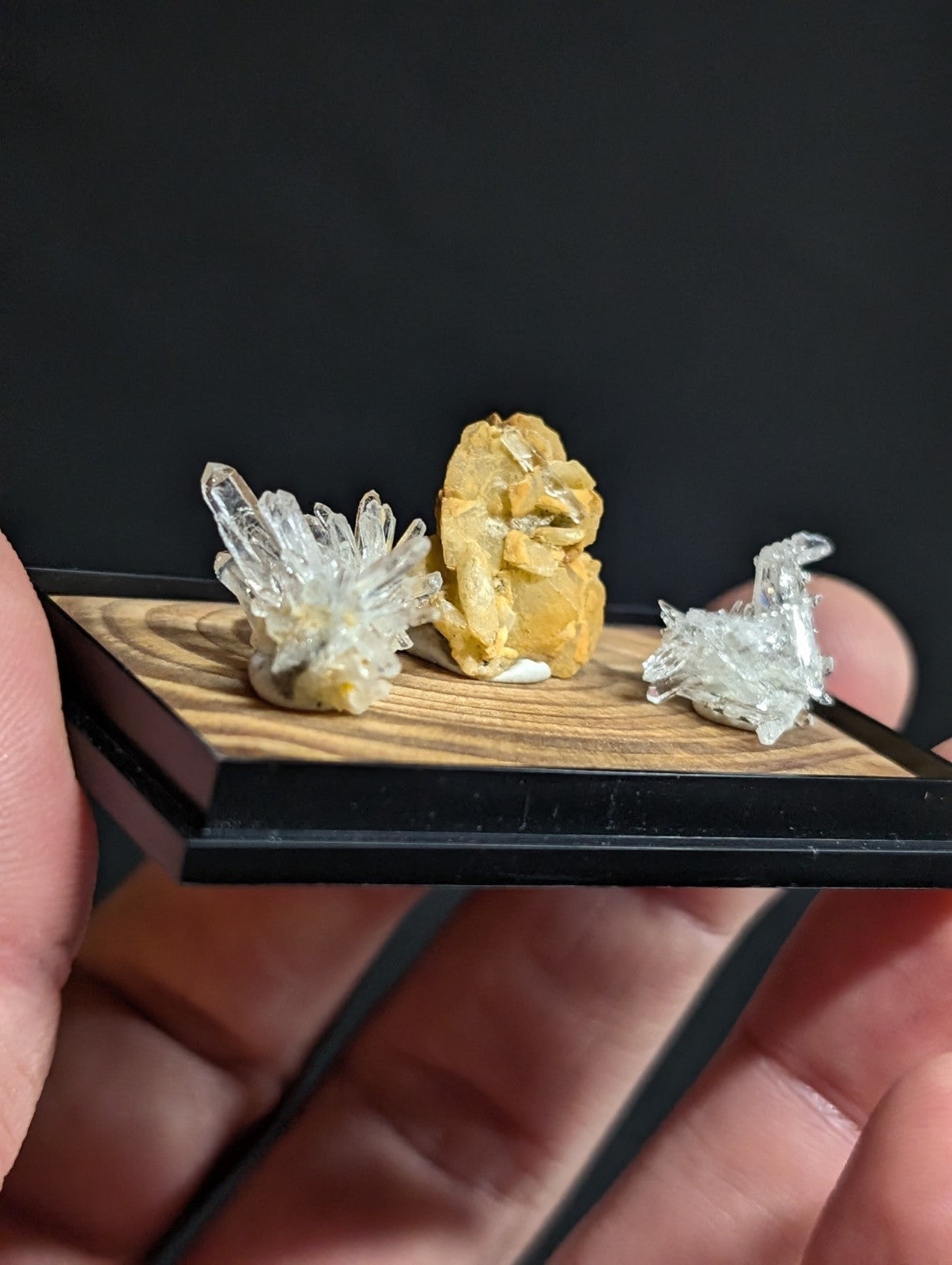 A miniature display of three crystal specimens—transparent quartz with sharp facets, a golden-yellow mineral cluster, and another clear crystalline formation—all mounted on a wooden base within a black frame.