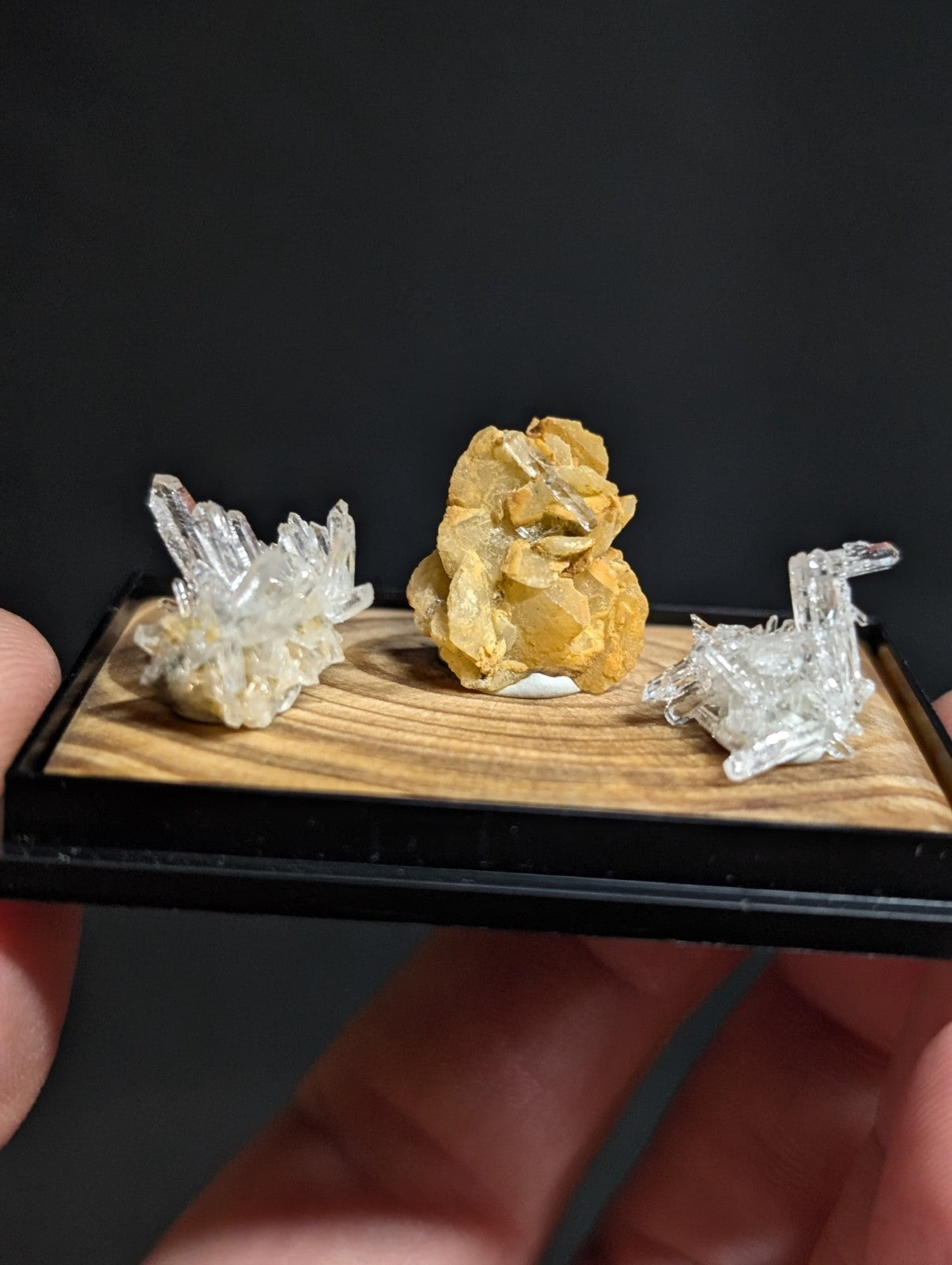 A miniature display tray holding three distinct mineral specimens: a cluster of clear quartz crystals, an orange-yellow geode with visible internal crystalline structure, and another transparent crystal formation.