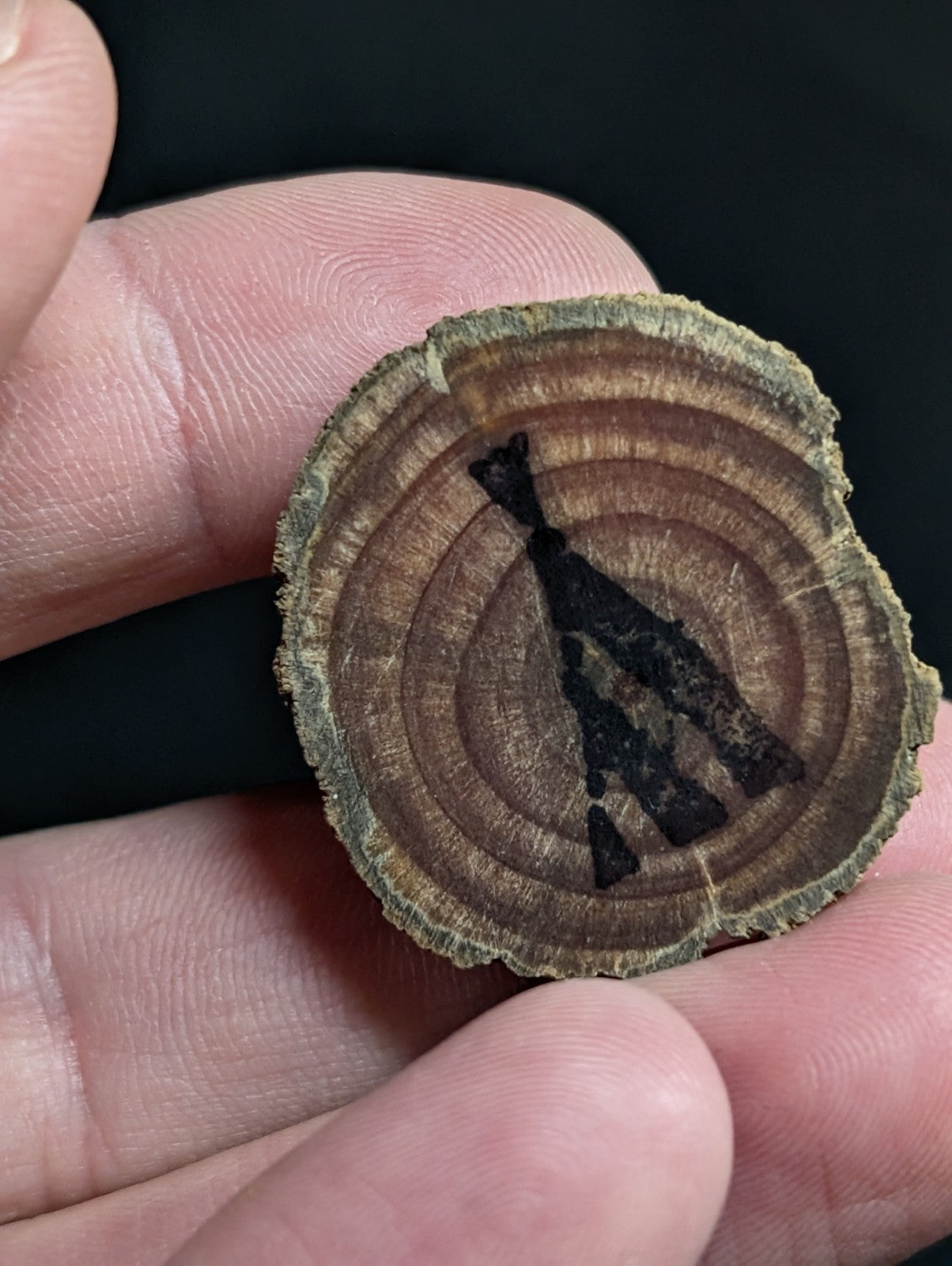 A circular, rustic wooden slice with visible growth rings and a dark charcoal-colored silhouette of a figure engraved on its surface.