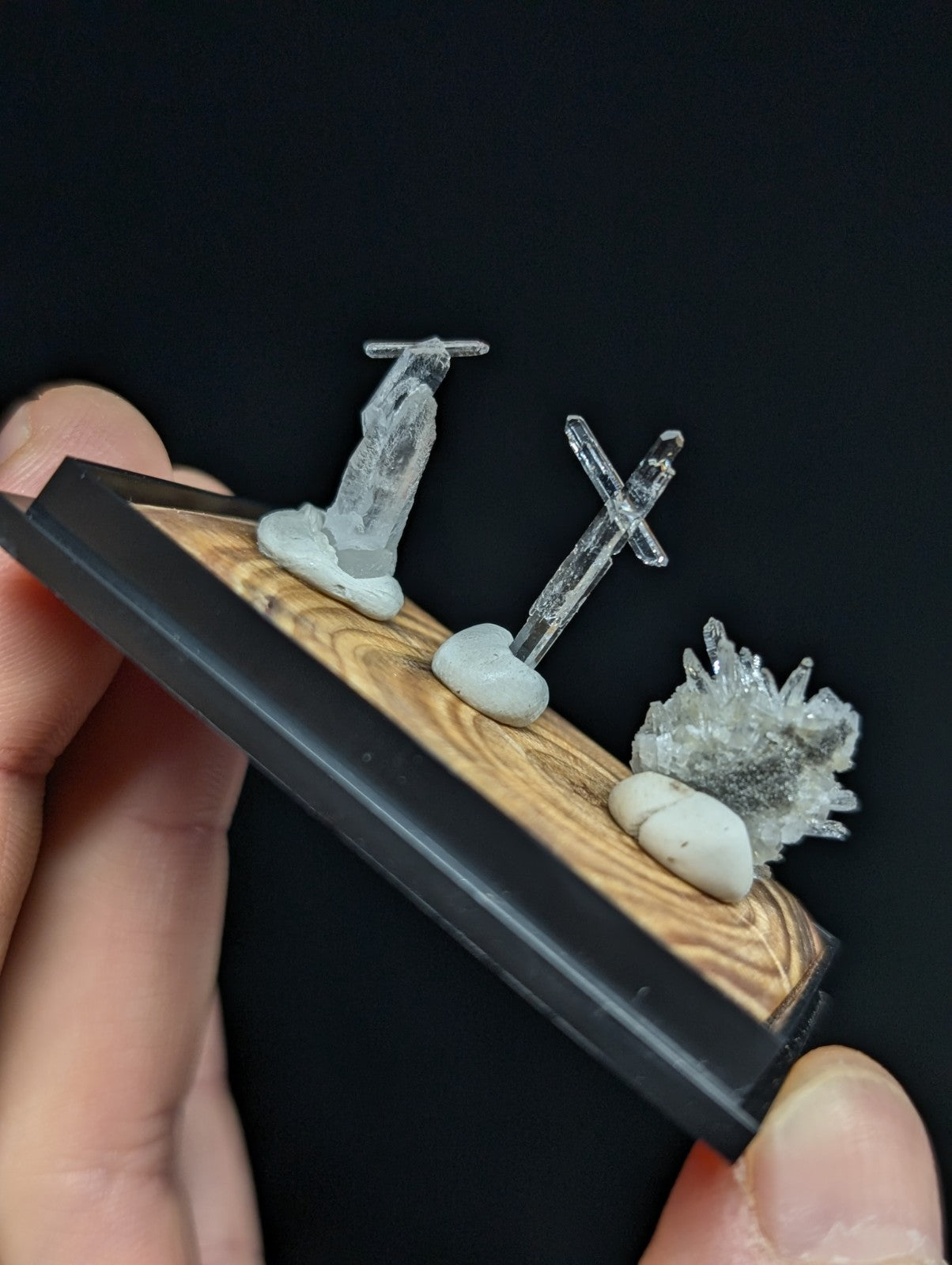 A miniature diorama featuring a translucent crystal statue, a clear cross-shaped ornament, and a cluster of sparkling crystals mounted on a wooden base with black trim.