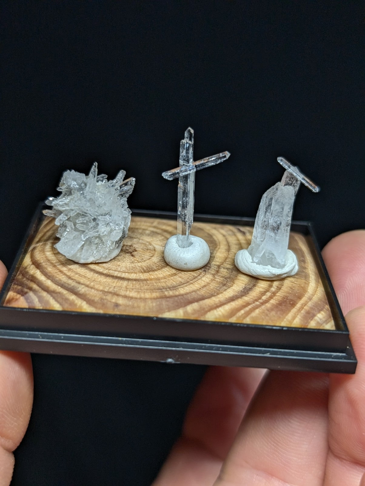 A clear quartz crystal display featuring a heart-shaped cluster, a cross, and a figure of the virgin mary, all mounted on a wooden base with black framing.