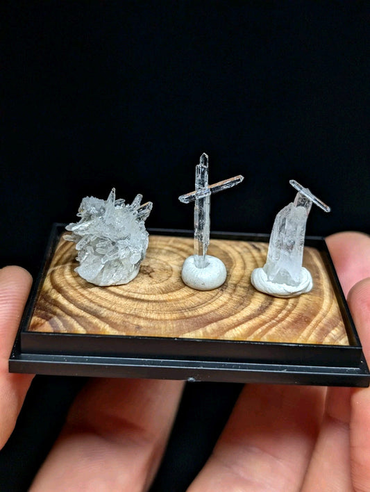 A clear quartz crystal display set featuring a cluster, a cross-shaped formation, and a standing figure—all mounted on a wooden base within a black frame.