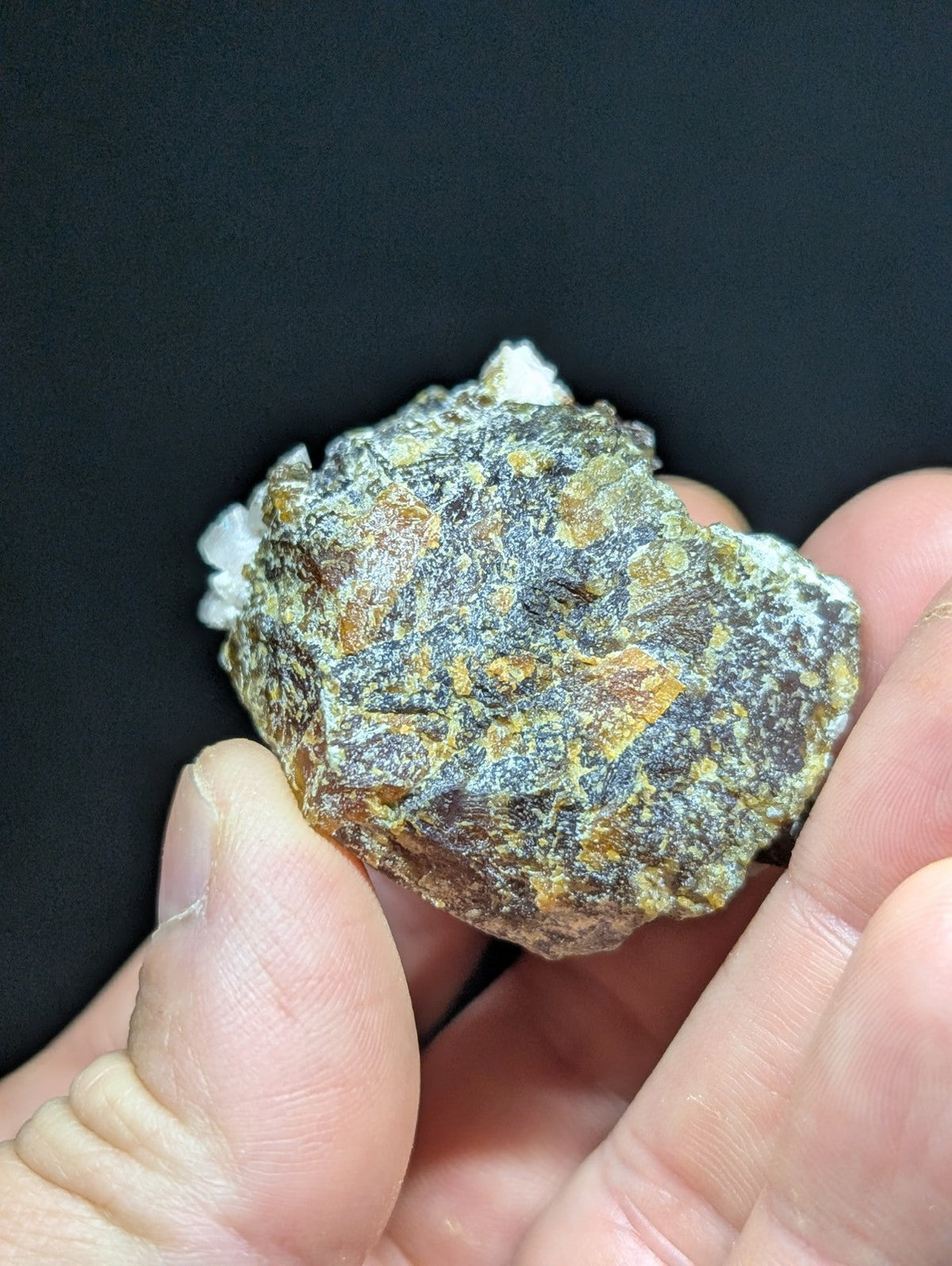 A rough, irregularly shaped mineral specimen with a mottled surface of dark gray, brown, and orange hues, accented by patches of white crystalline growths.