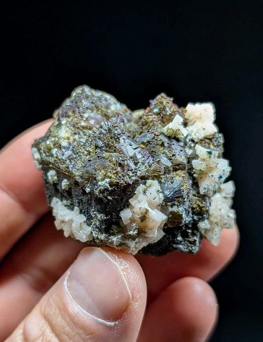 A hand holds a rough, dark green and black mineral specimen with prominent white crystalline formations.