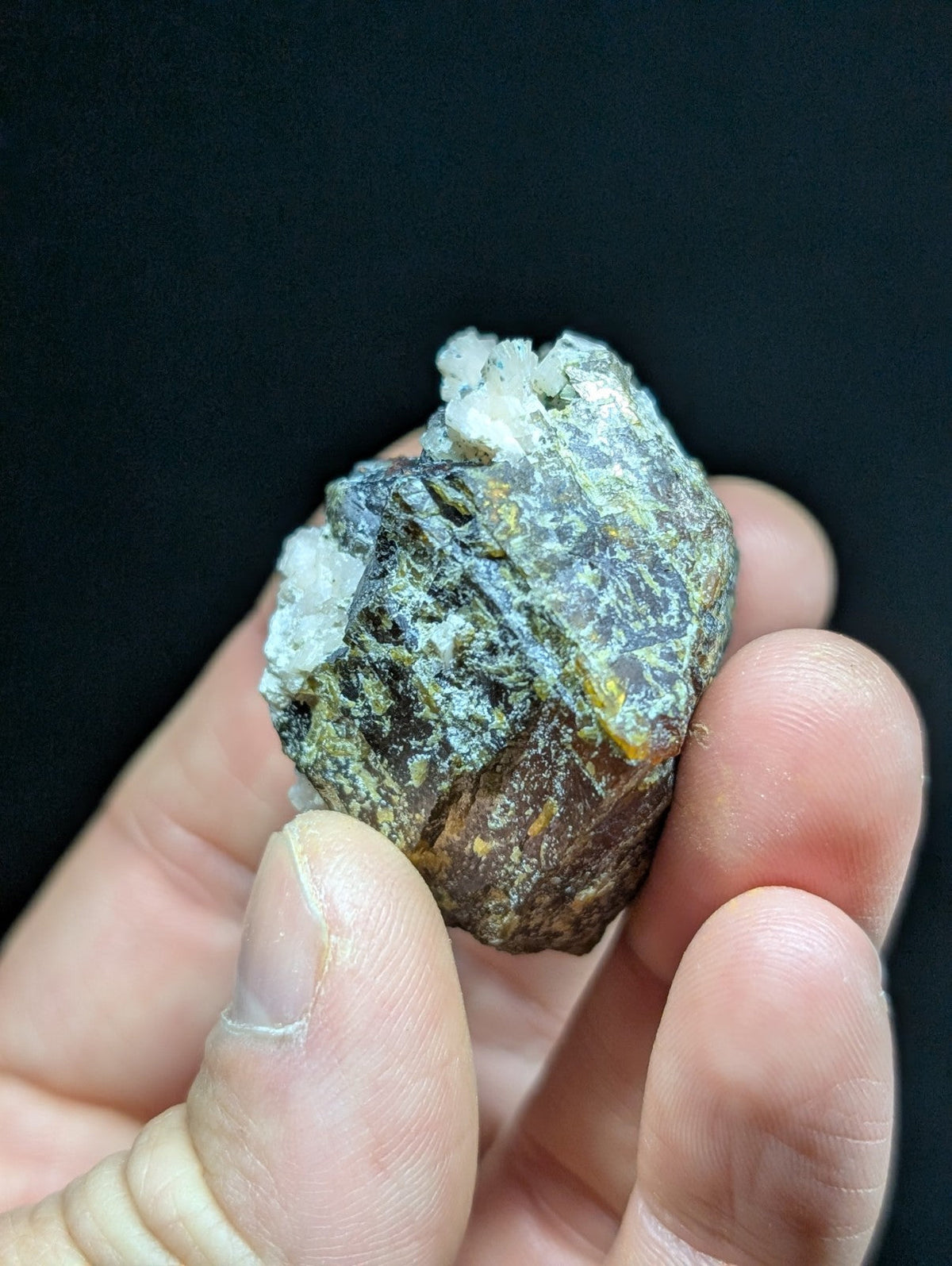 A rough, irregularly shaped mineral specimen with a mottled surface of dark brown, greenish-gray, and patches of white crystalline material.
