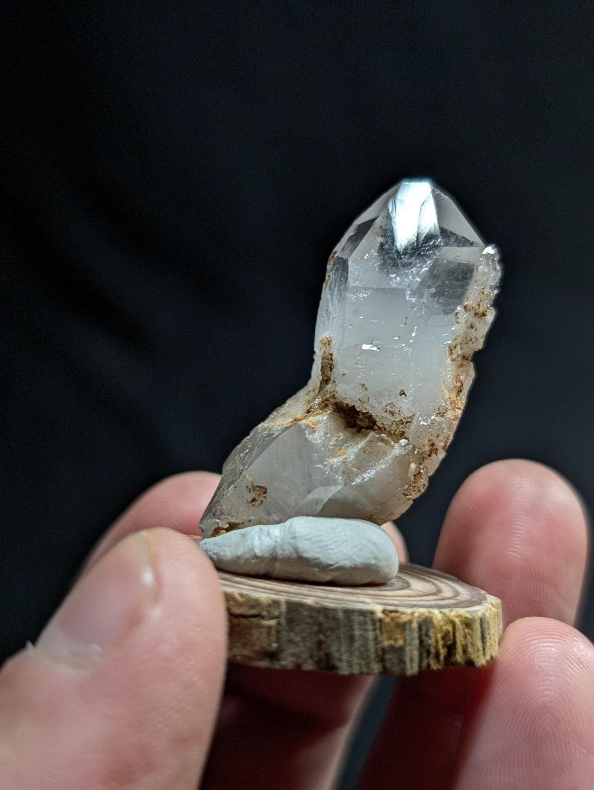 A clear quartz crystal with a pointed tip and natural matrix, displayed on a small wooden slice.