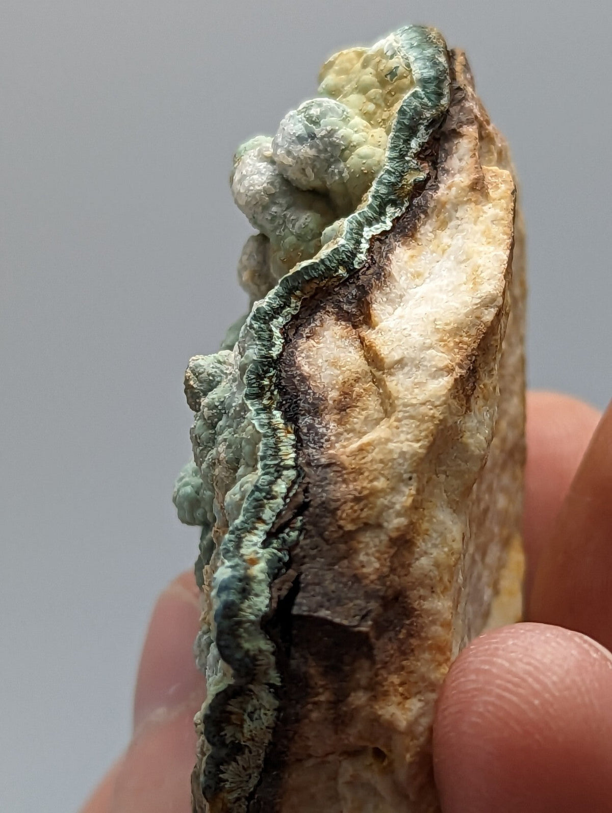 A hand holds a rough, irregularly shaped mineral specimen featuring a striking greenish-blue veining against a tan and brown matrix.
