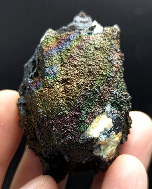 A rugged, irregularly shaped meteorite with a striking iridescent surface displaying shimmering hues of green, gold, and purple against its dark, textured matrix.