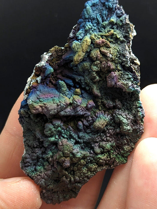 A hand holds a rugged, iridescent mineral specimen with swirling patterns of deep blue, green, purple, and gold.