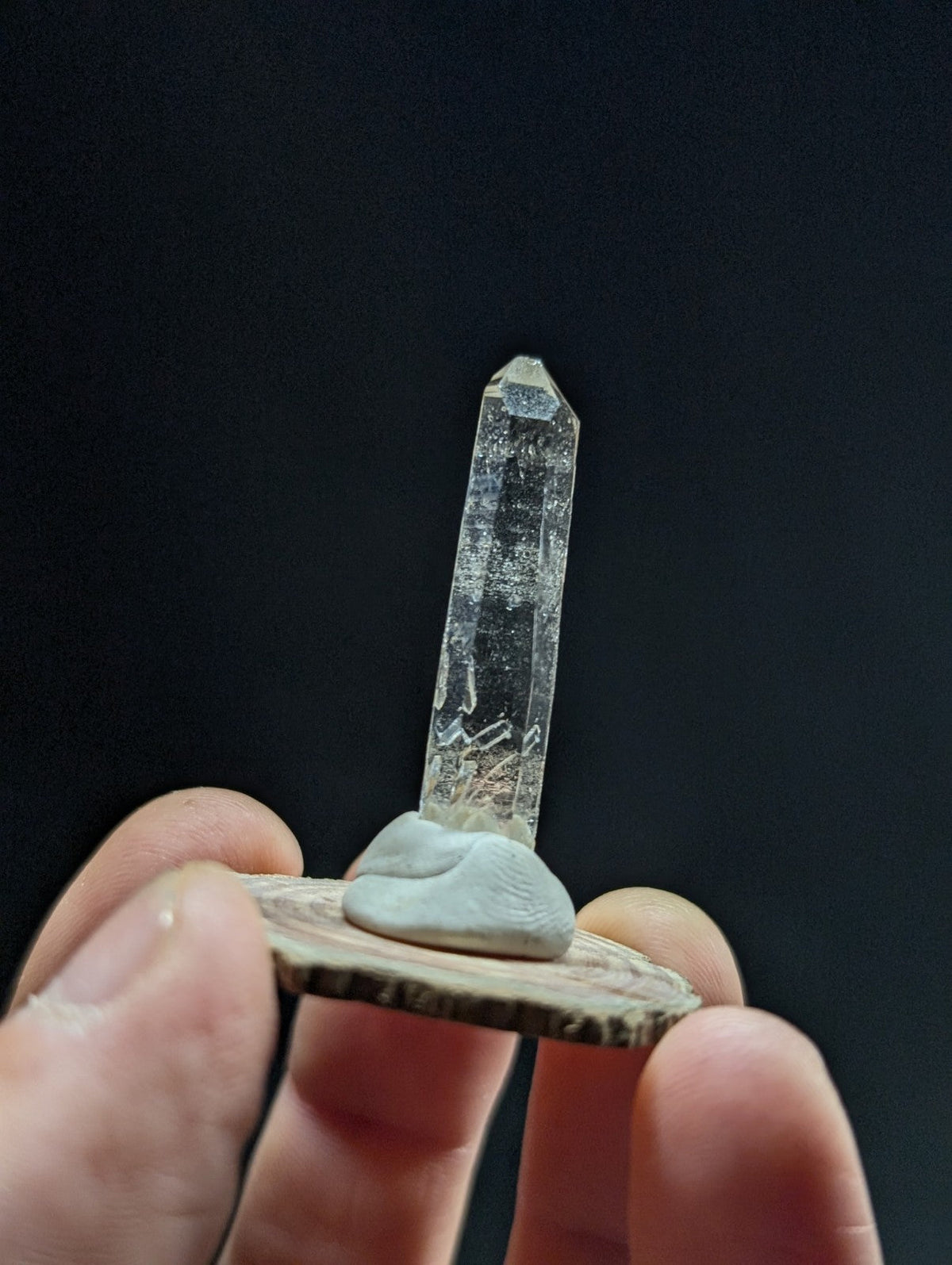 A clear, faceted quartz crystal with a pointed tip and natural inclusions stands upright on a small, light-gray clay base mounted on a circular wooden slice.