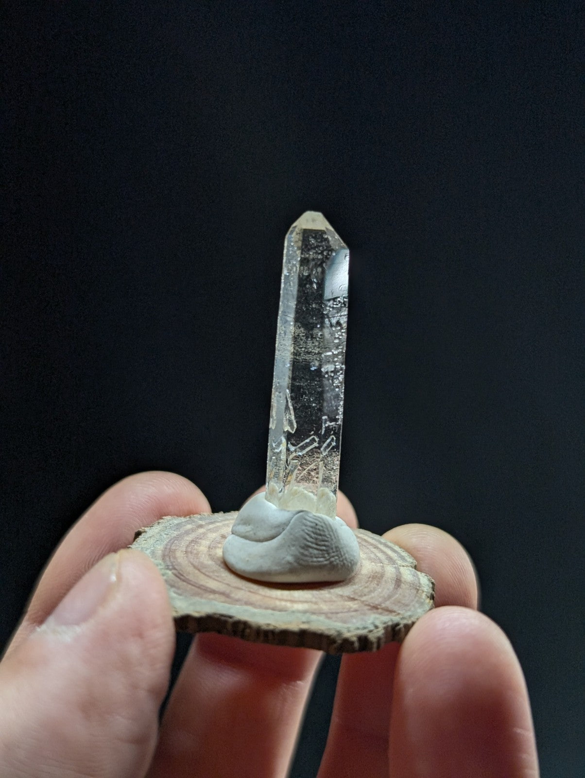 A clear quartz crystal with a pointed tip, mounted on a light gray stone base that sits atop a circular wooden slice.