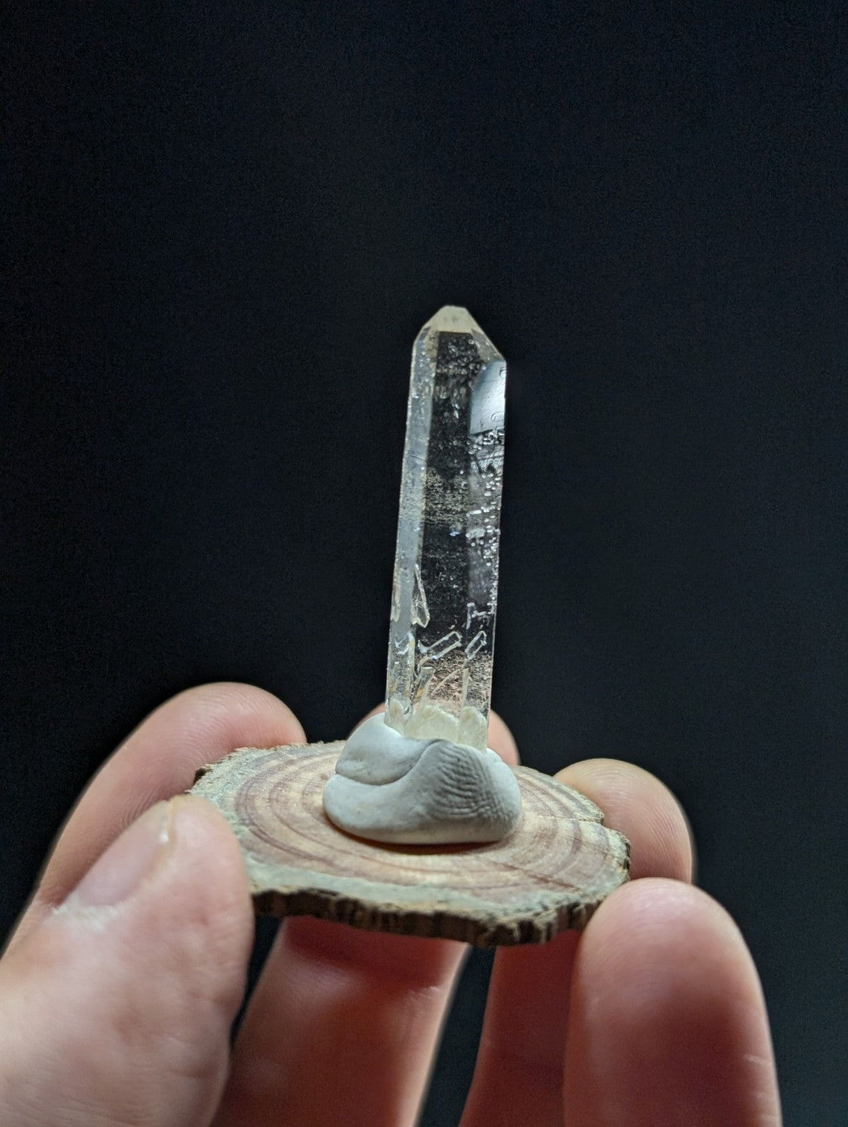 A clear quartz crystal with a pointed tip, mounted on a light gray stone base that sits atop a circular wooden slice.