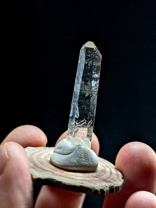 A clear, translucent quartz crystal with a pointed tip and natural inclusions stands upright on a small, grayish stone base set within a circular wooden slice.
