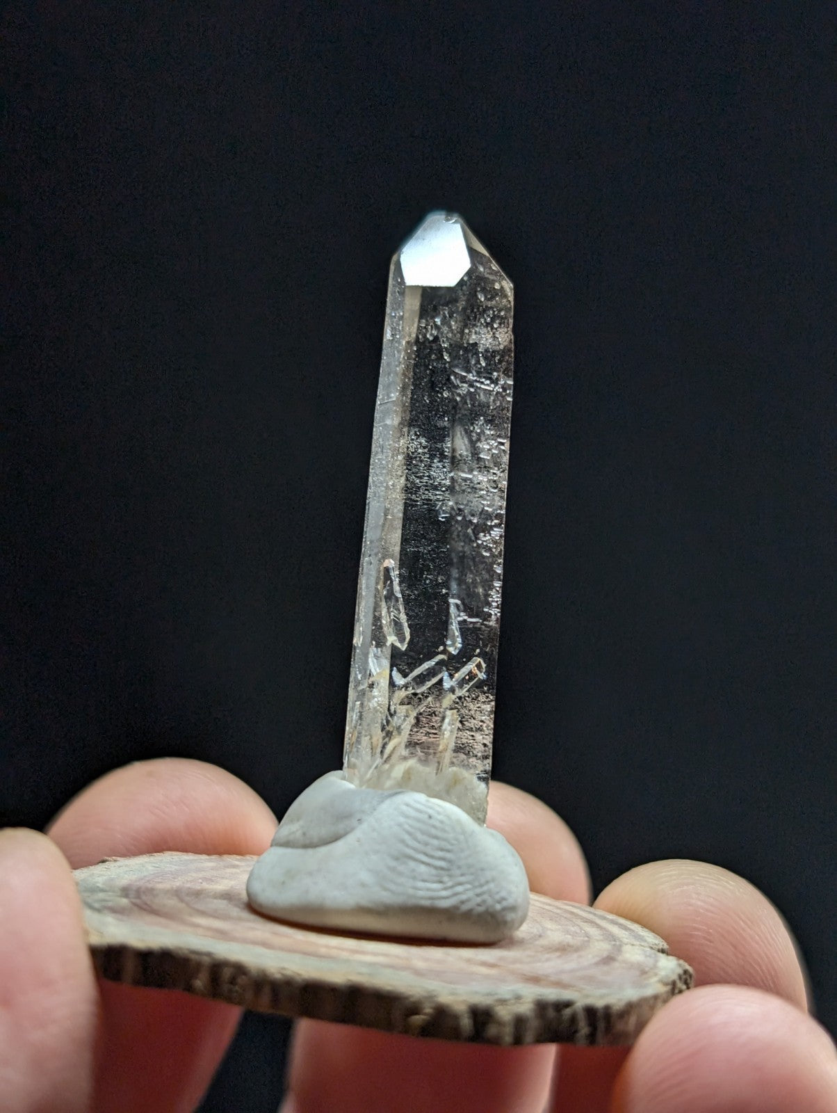 A clear quartz crystal with a pointed tip and natural inclusions, displayed upright on a smooth gray stone resting atop a circular wooden slice.