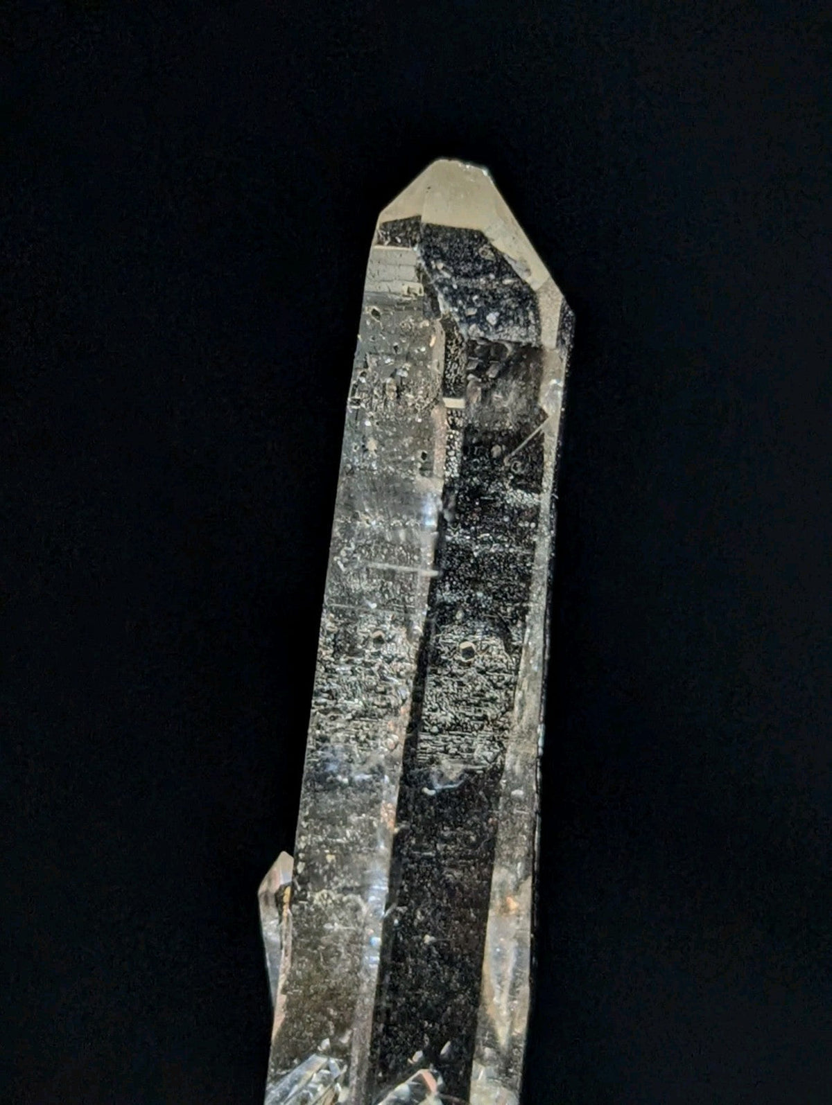 A translucent quartz crystal with a pointed tip and internal dark inclusions, featuring natural striations and subtle imperfections.