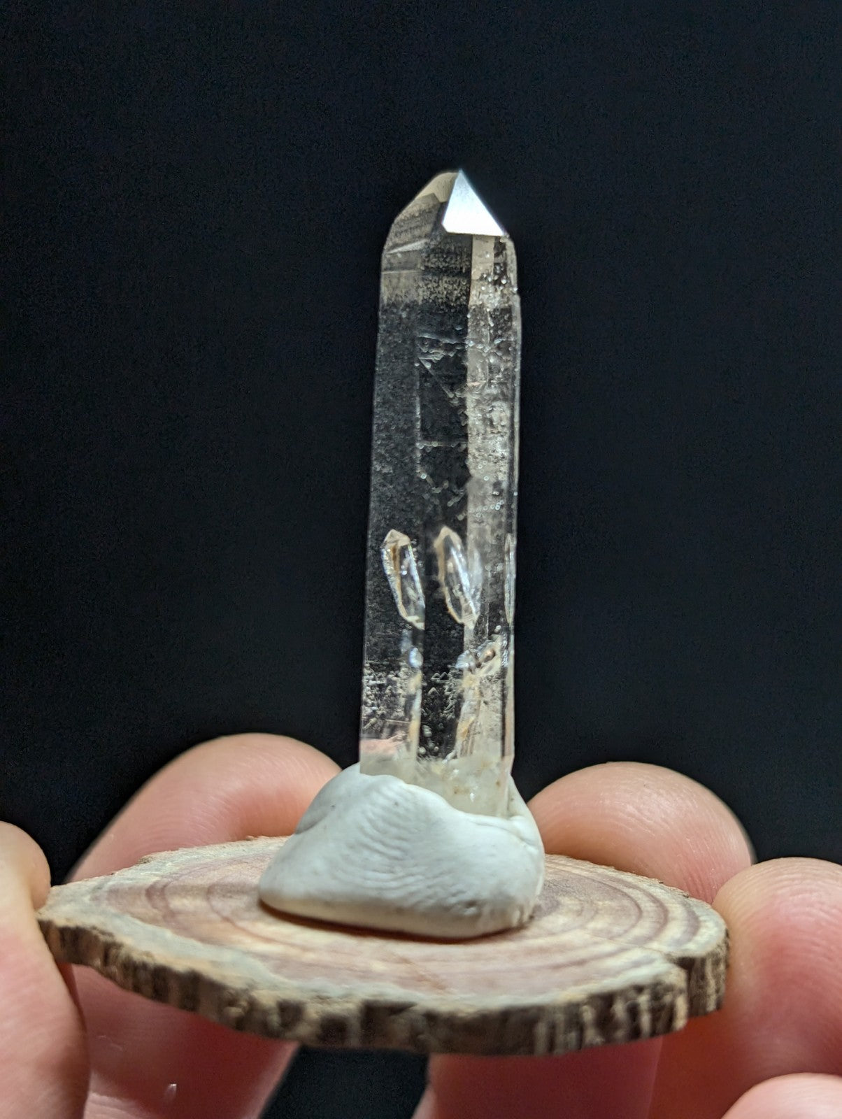 A clear quartz crystal with a pointed tip and natural inclusions, mounted on a rustic wooden slice base.
