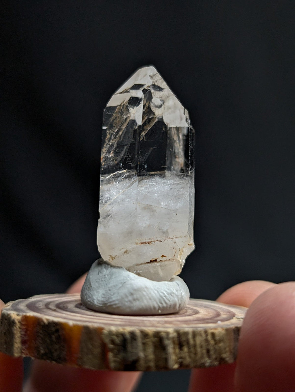 A clear, faceted quartz crystal with a translucent body and subtle internal inclusions sits atop a smooth, light-gray stone base.