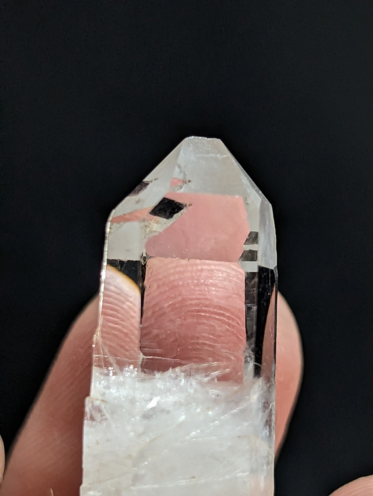 A clear quartz crystal with a pinkish inclusion and visible internal fractures, held between fingers against a dark background.