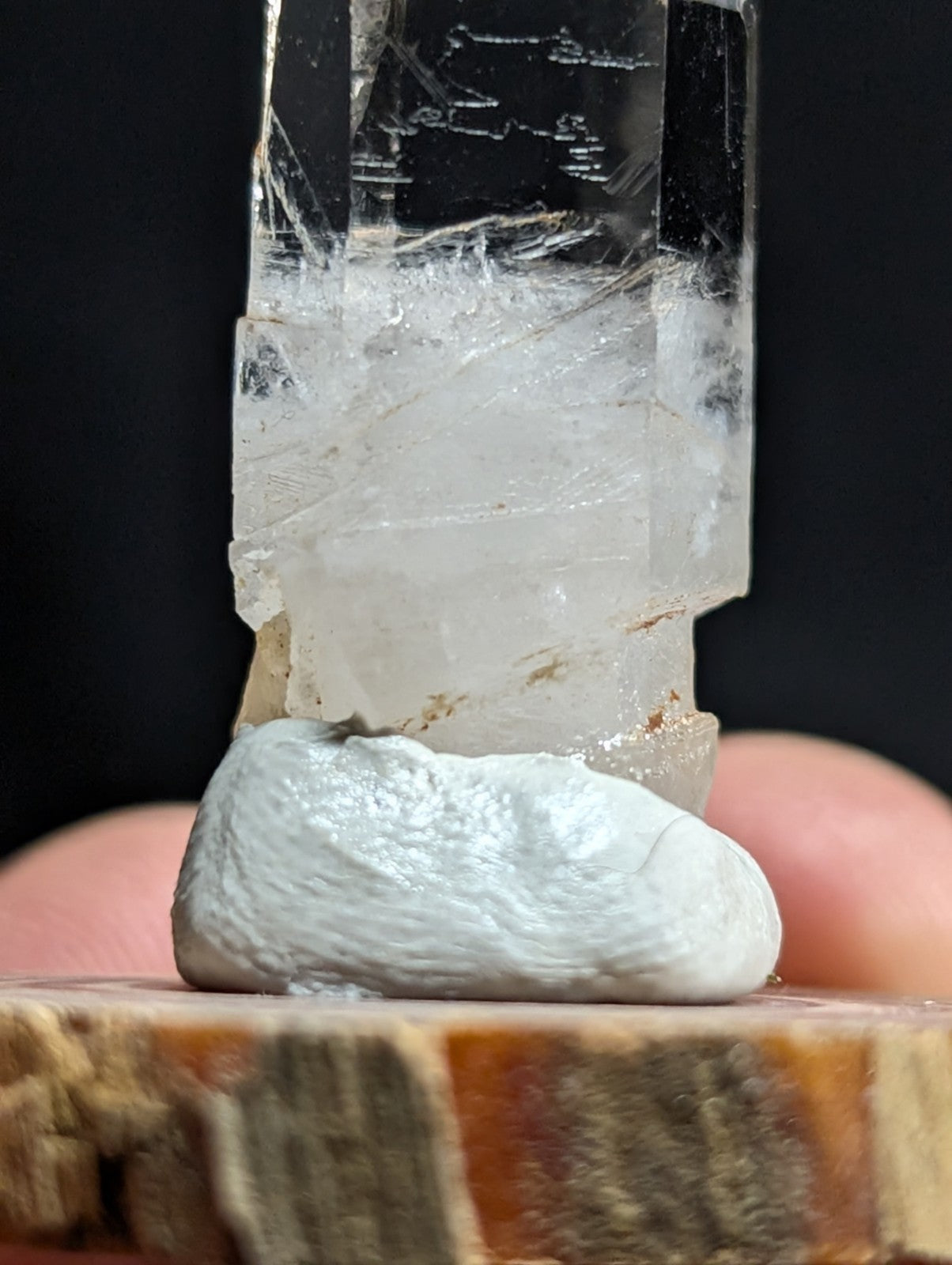 A clear, translucent quartz crystal with visible internal striations and a rough base rests on a smooth white stone atop a wooden surface.