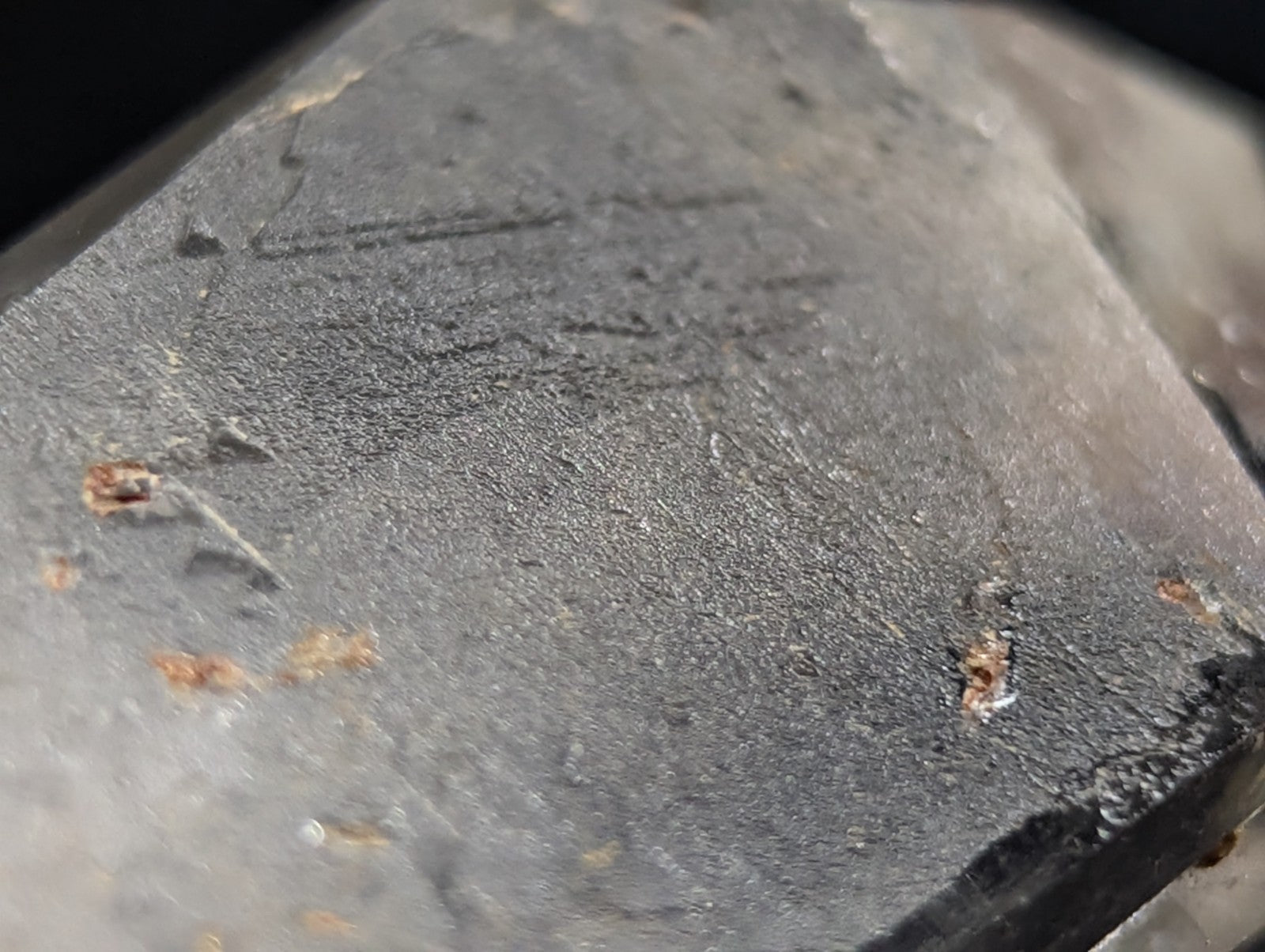 A dark gray, rough-textured mineral crystal with subtle surface markings and small brownish inclusions.