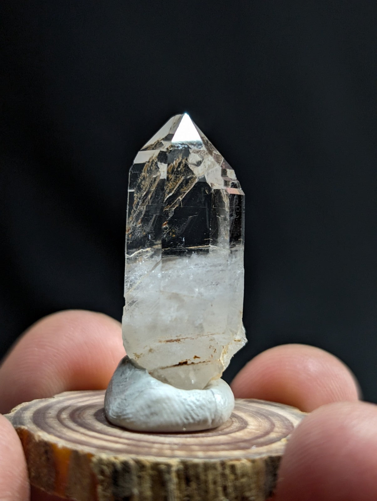 A clear, faceted quartz crystal with natural inclusions and a pointed tip, displayed on a small white pedestal atop a wooden slice.