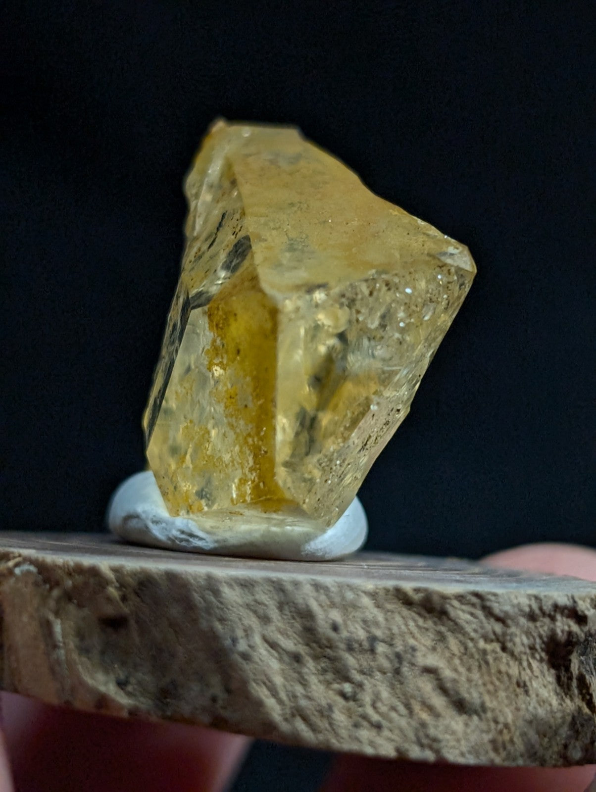 A translucent yellow gemstone with a rough, faceted surface and internal inclusions rests on a white pedestal atop a textured stone slab.