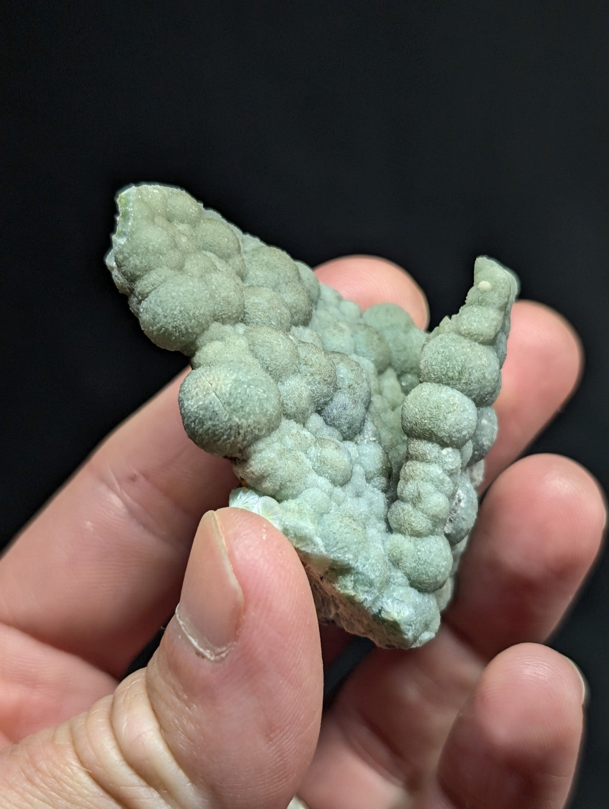 A hand holds a light green, textured mineral specimen with rounded, bubble-like formations.