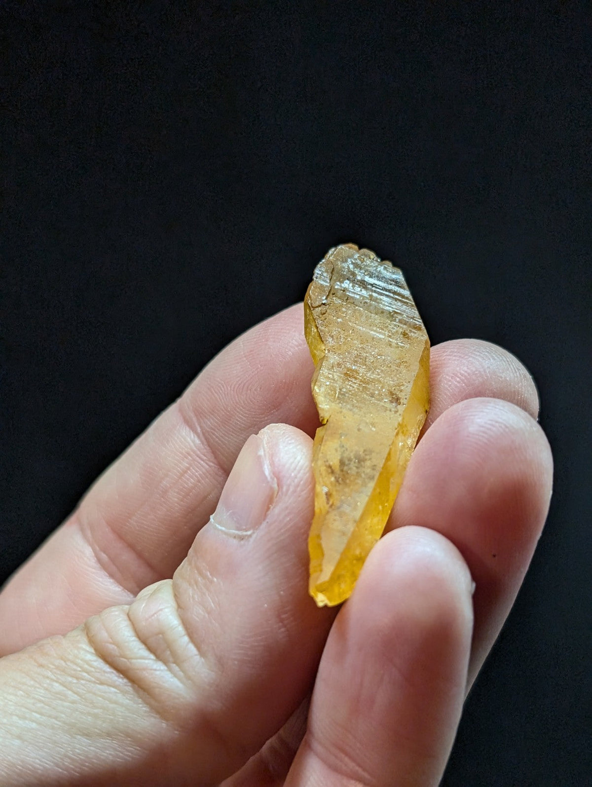 A translucent yellow crystal with a pointed tip and striated surface, held gently in someone’s hand.