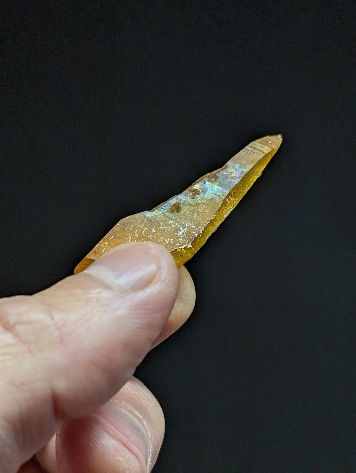 A translucent amber-colored crystal with a pointed tip and subtle iridescent flashes, held delicately between fingers.