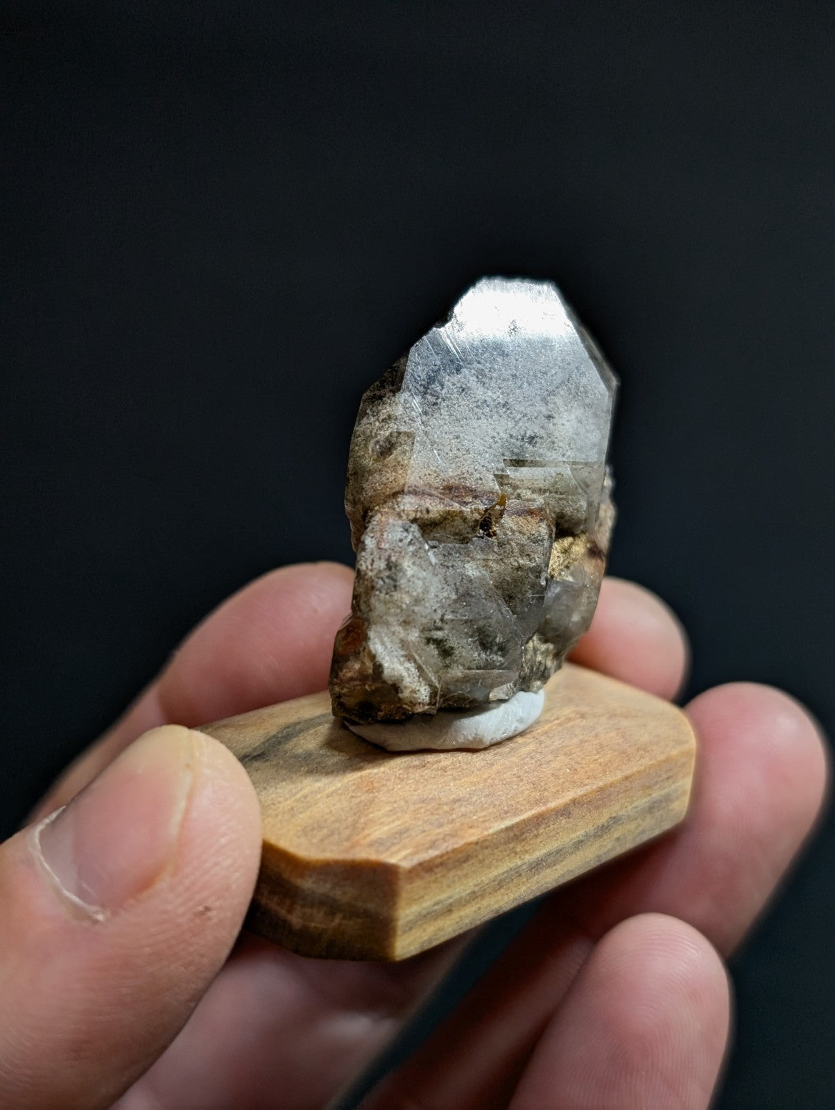 A polished, translucent quartz crystal with a metallic sheen and earthy brown veining is displayed on a small wooden stand.