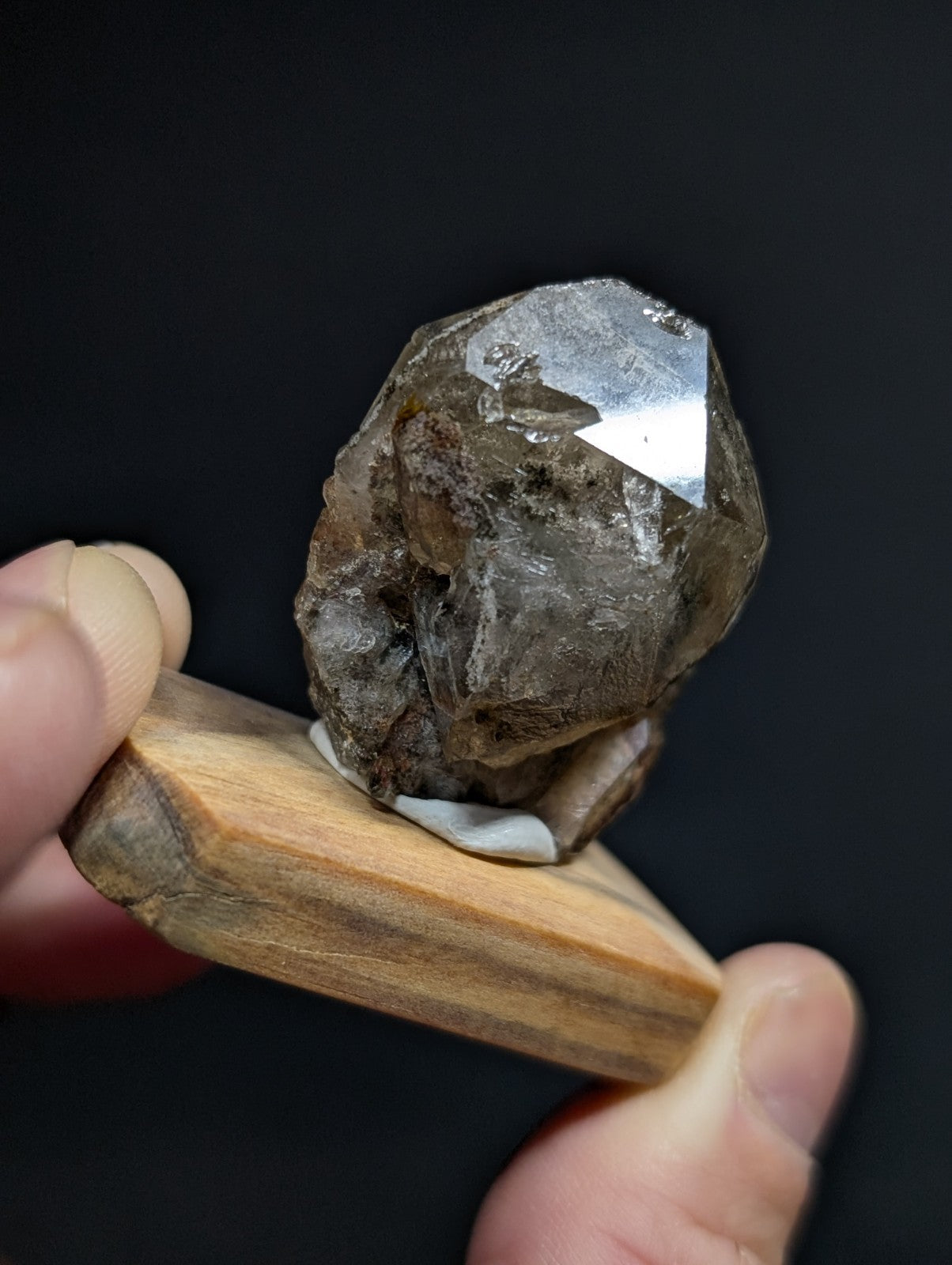 A rough, translucent smoky quartz crystal with a faceted top and earthy brownish-gray tones, held in place by wooden display stand.