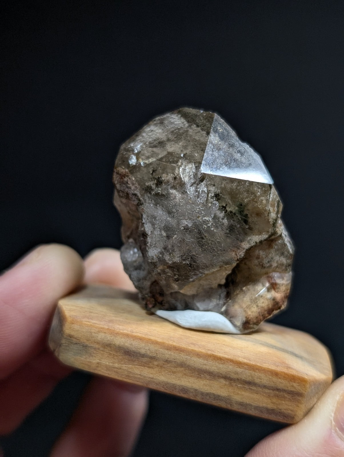 A rough, translucent smoky quartz crystal with a prominent clear facet and earthy brownish-gray tones, displayed on a wooden stand.