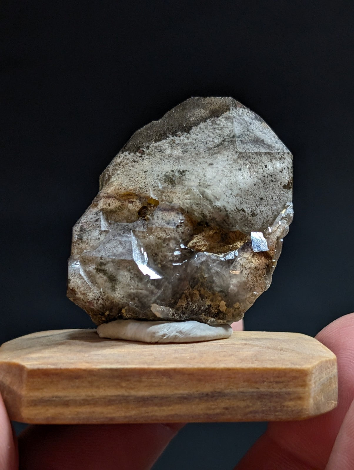 A rough, translucent quartz crystal with gray and brown veining rests on a light-colored wooden display stand.