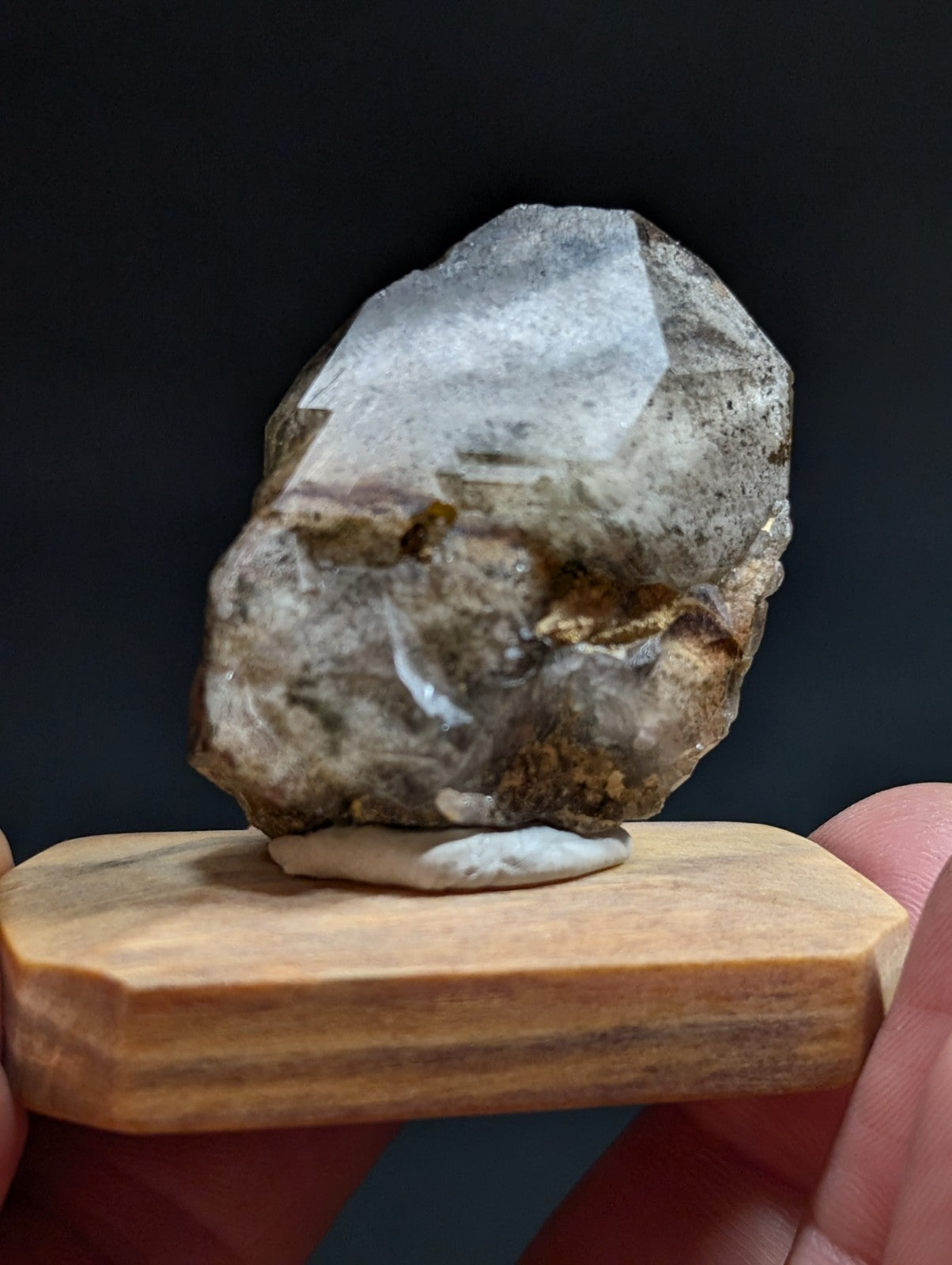 A translucent, grayish-white crystal with natural inclusions and a rough, irregular surface rests on a wooden display stand.