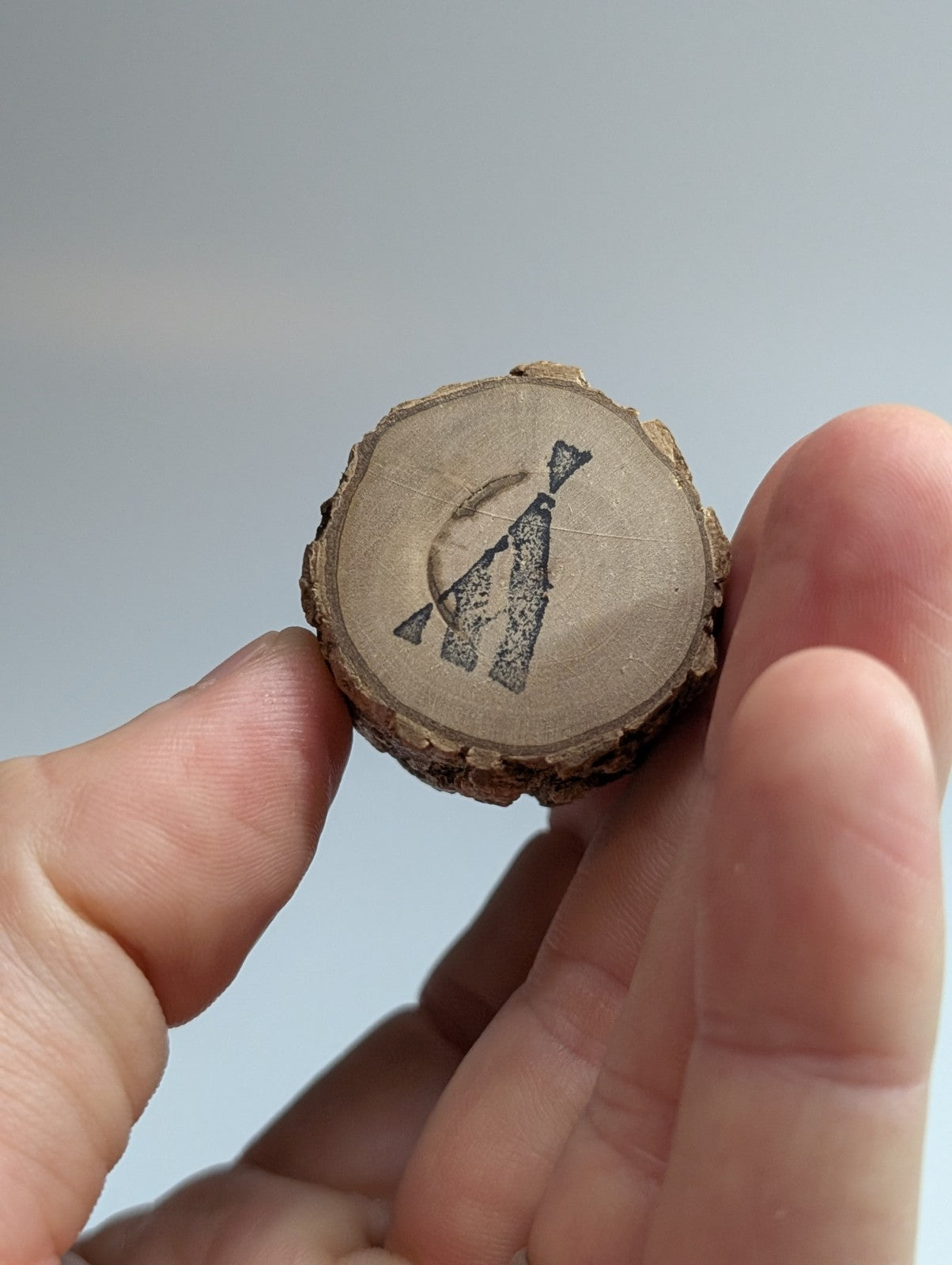 A circular, rustic wooden slice with a natural bark edge and a dark charcoal engraving of an abstract figure.