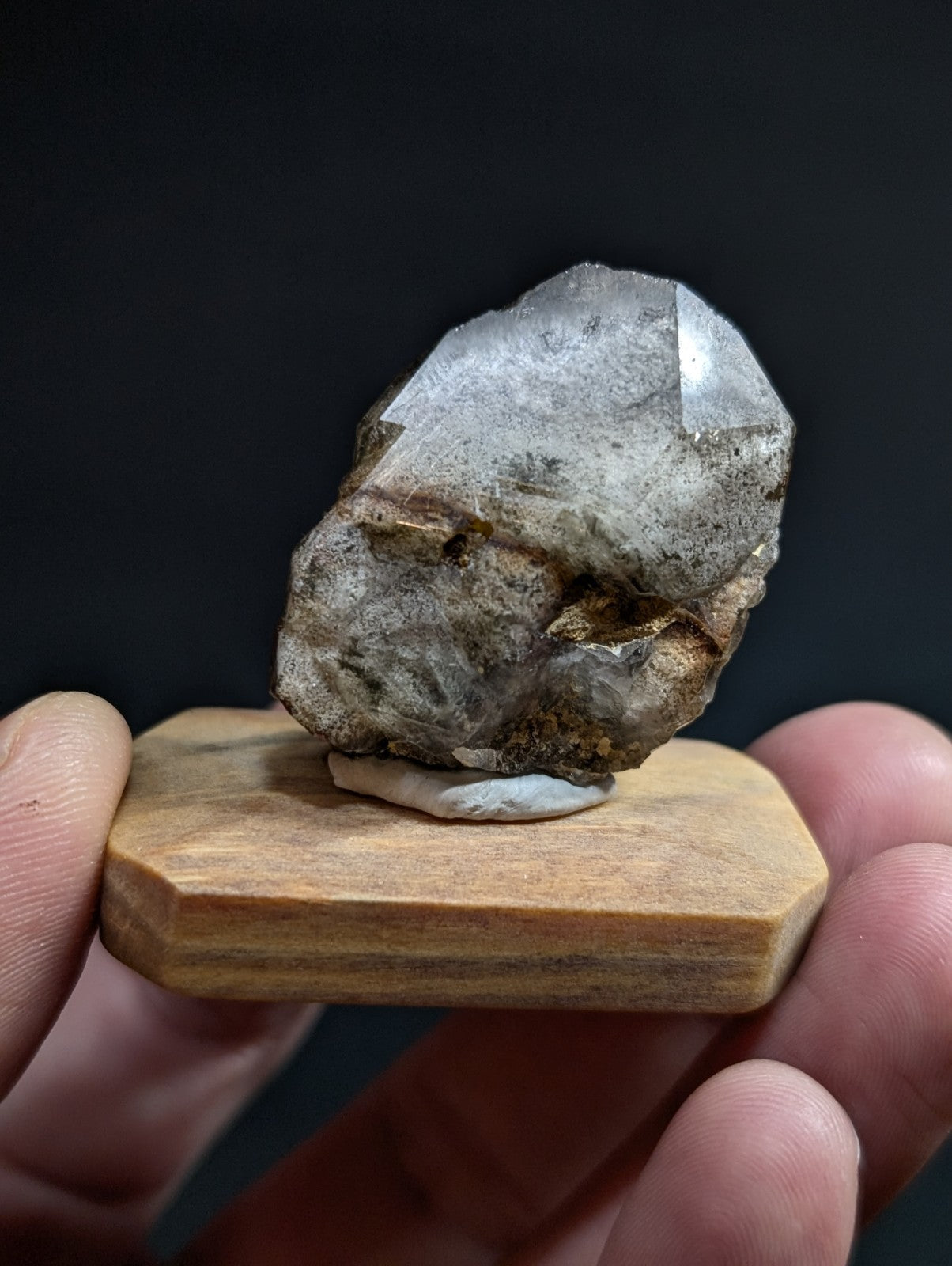 A translucent, grayish-white quartz crystal with a rough, earthy base and sharp facets sits atop a light-colored wooden display stand.